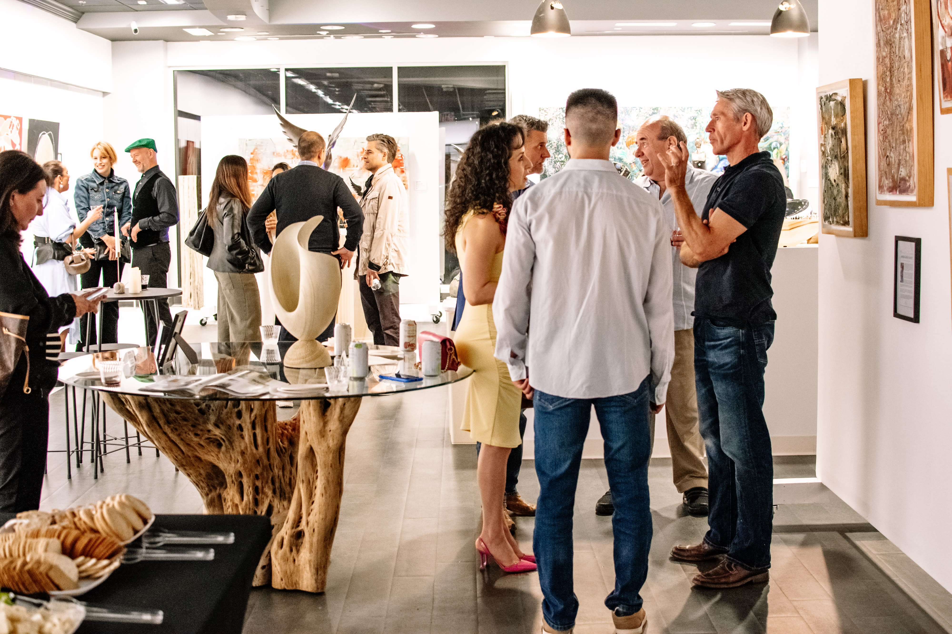 Guests mingle and socialize at a birthday party held in a spacious indoor venue, with a variety of artworks displayed on the walls and tables set for refreshments.