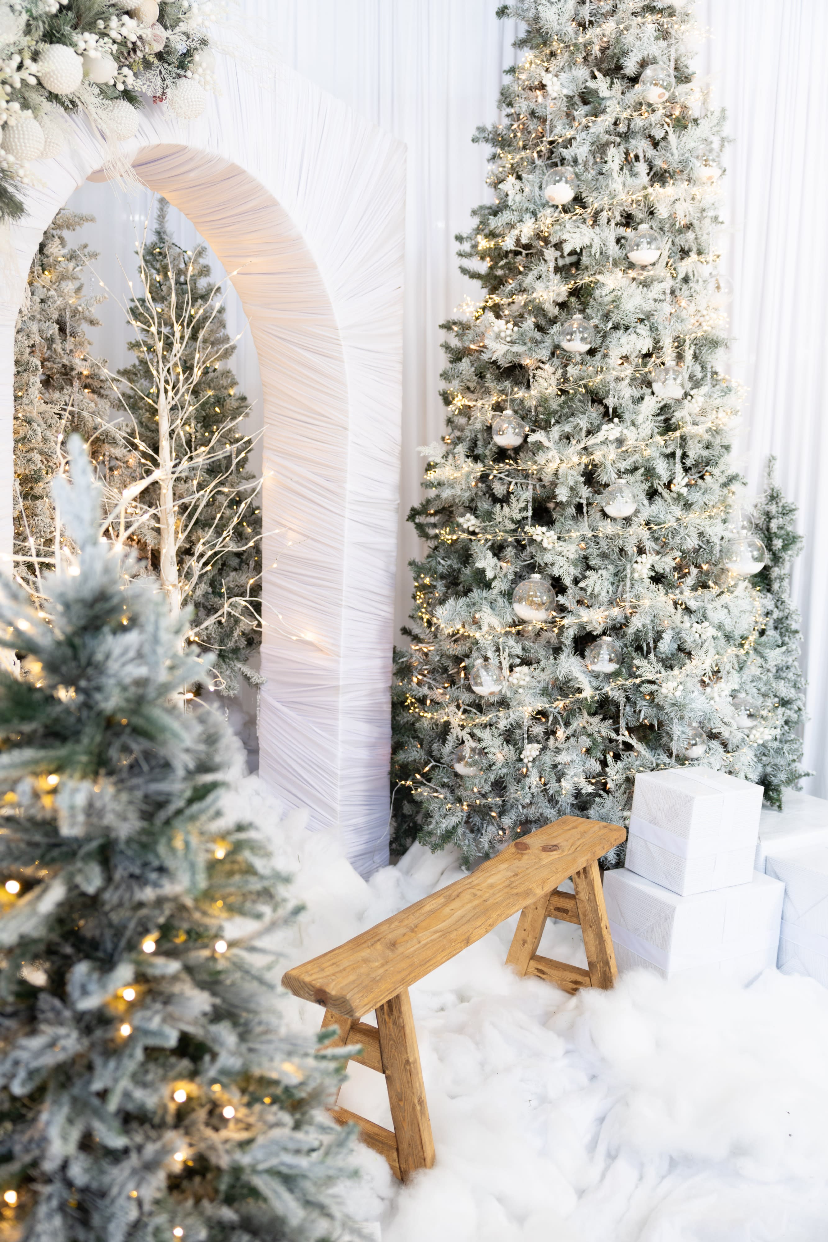 A child in a black dress sits on a light-colored chair in front of a decorated Christmas tree and gift boxes, all set against a red backdrop.