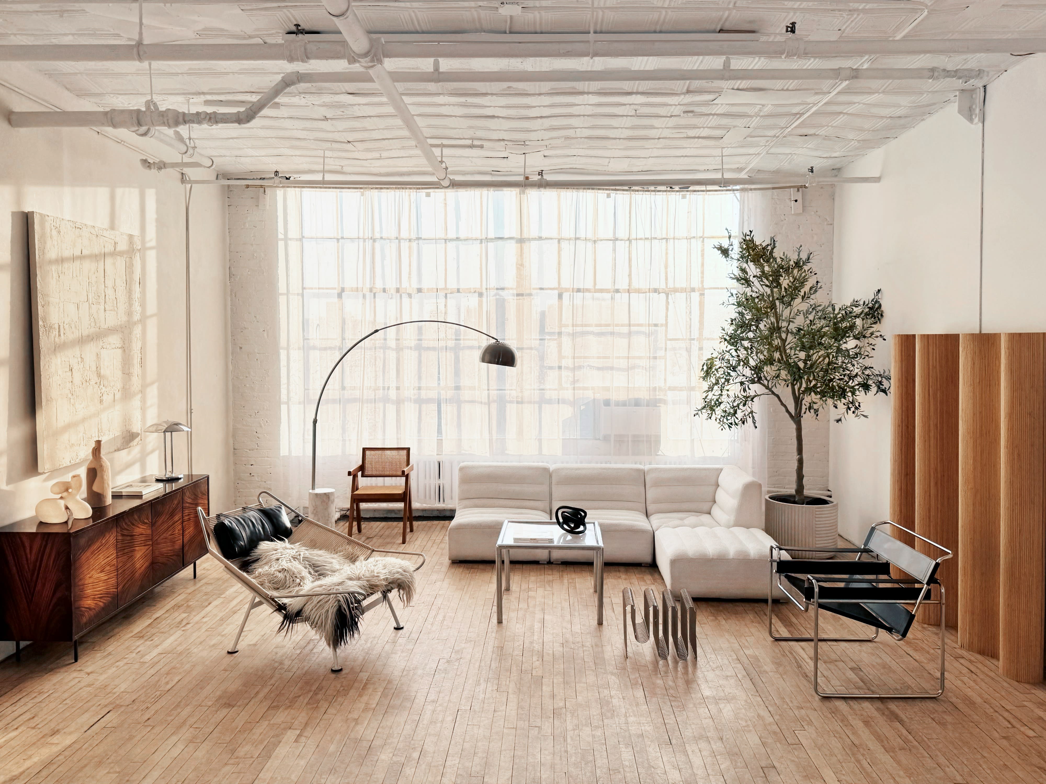 The photo shows a minimalist living area featuring a white sectional sofa, a glass coffee table, and a decorative plant beside a large window.