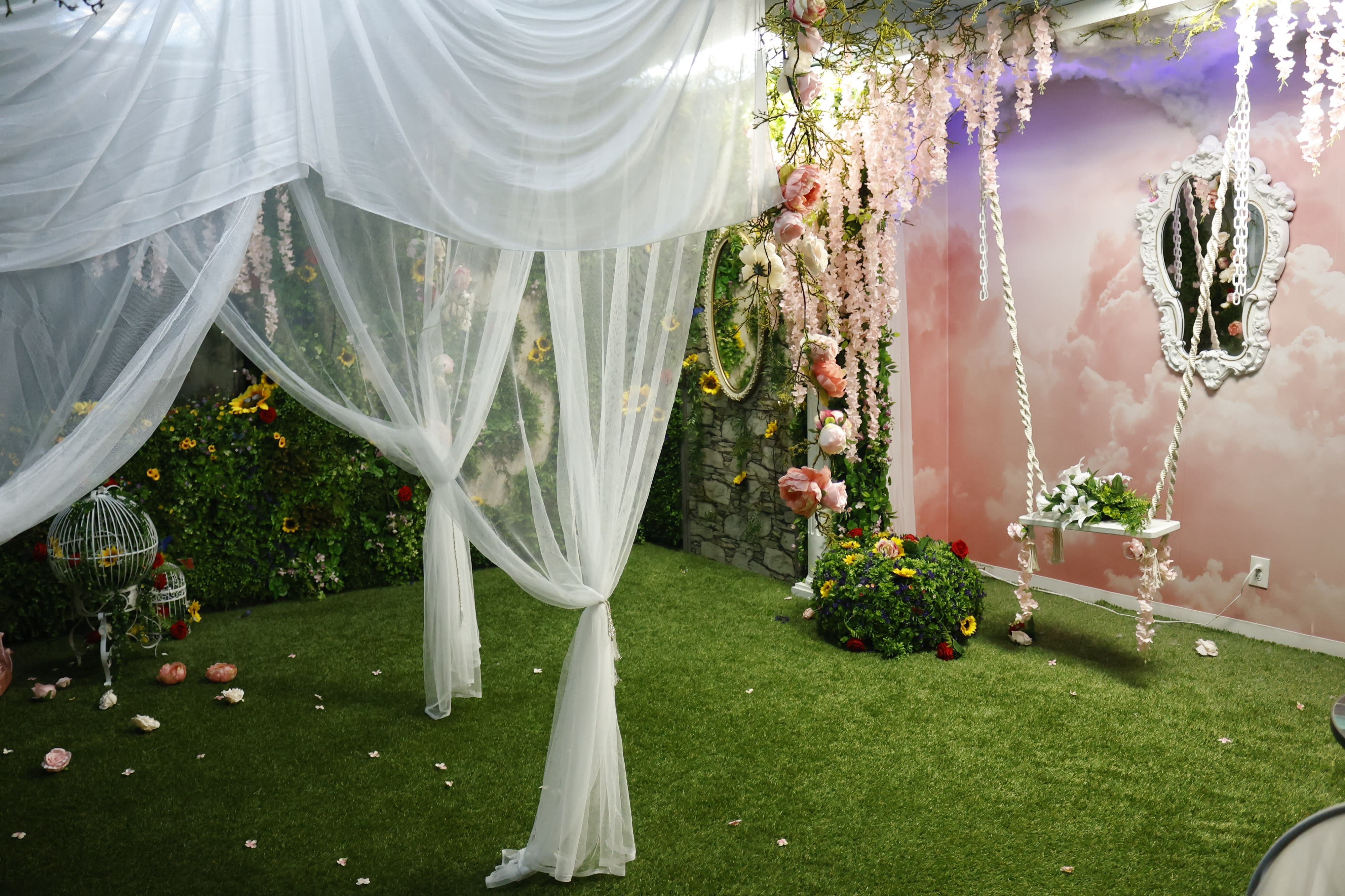 A young woman in a decorative dress sits on a swing surrounded by a pink cloud-themed backdrop and floral accents.