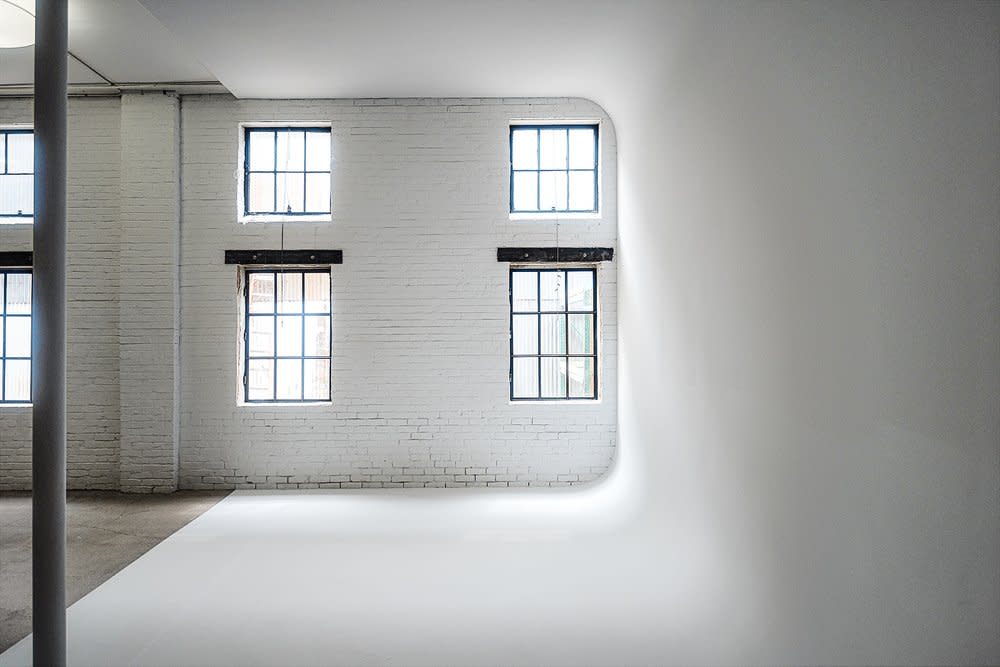 A model poses in a flowing white gown and cape made of floral fabric, leaning against a white brick wall near a large window.