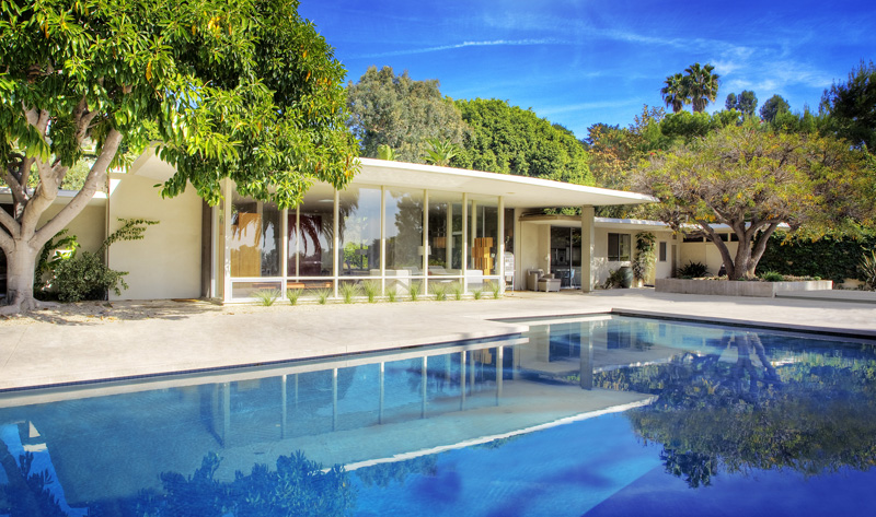 A filmmaker directs a scene by a pool in a modern backyard surrounded by lush greenery.