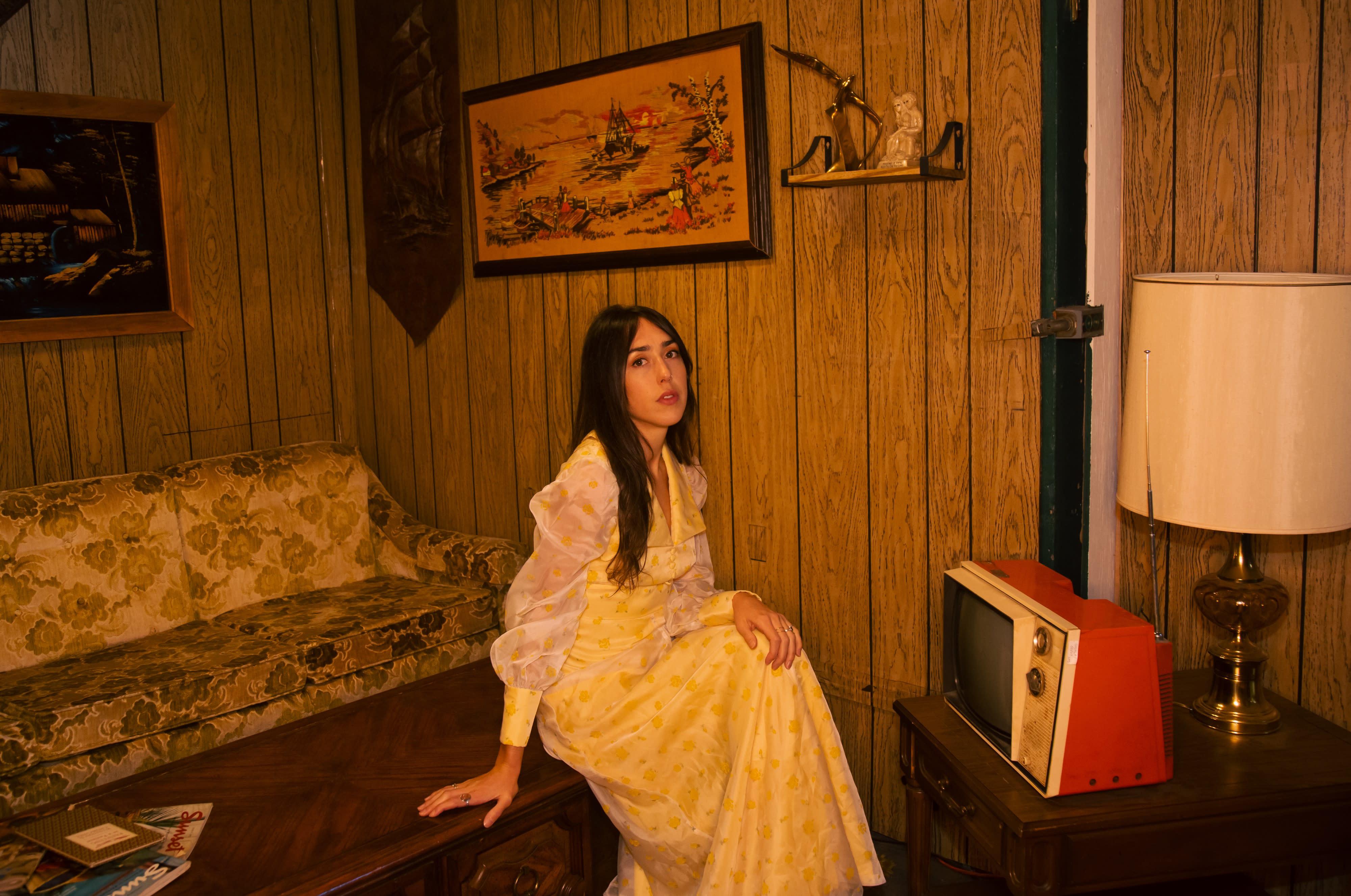 A band performs in a cozy, retro-styled living room, featuring acoustic and electric instruments.