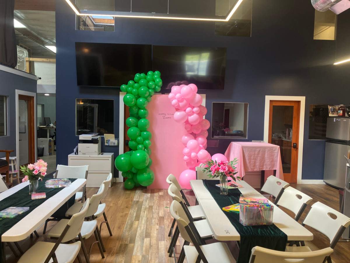 A group of guests gathers around tables at a venue, celebrating a 40th birthday with decorations, a large illuminated "40," and a projector screen displaying a presentation.