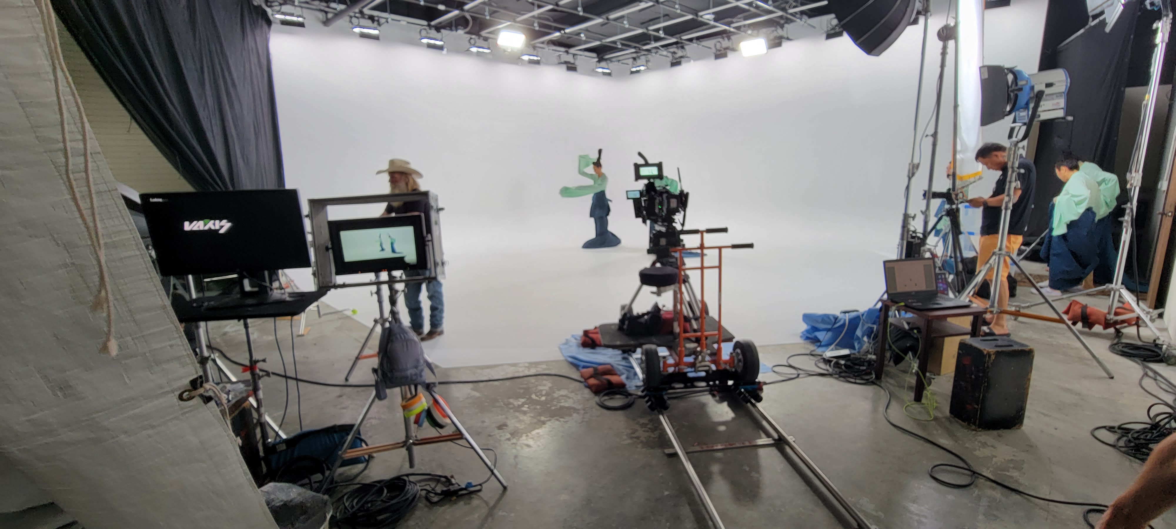 A photoshoot takes place in a studio with a backdrop, a camera setup, lighting equipment, and a ladder in the background.