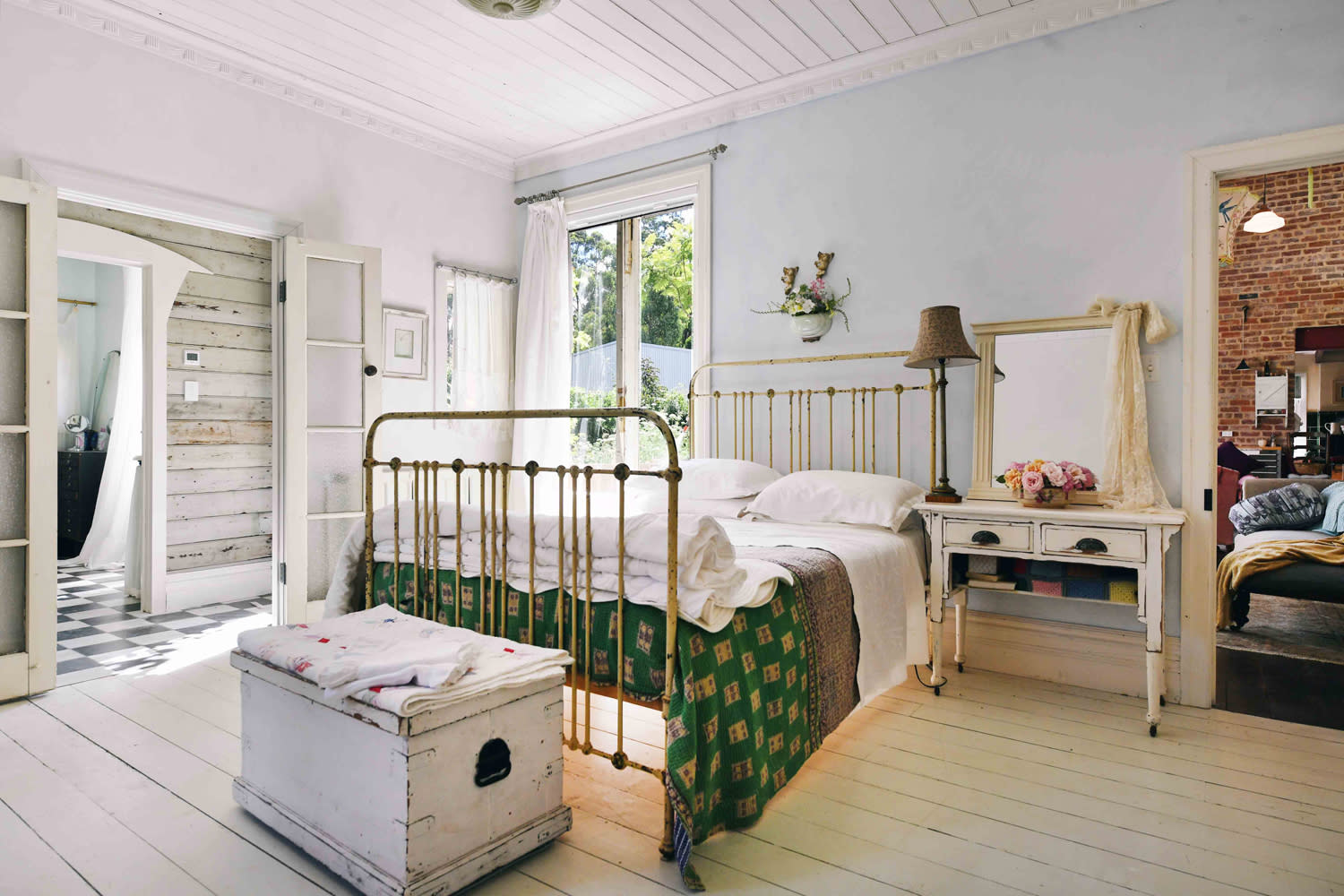 A film crew sets up equipment in a vintage-style bedroom featuring a metal bed frame, antique mirror, and soft natural lighting.