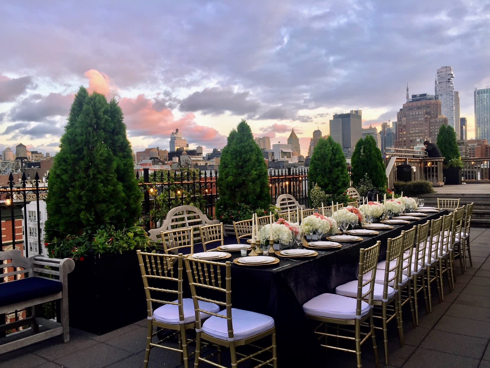 A couple is preparing for a romantic proposal on a rooftop decorated with candles and flowers, set against a skyline backdrop at night.