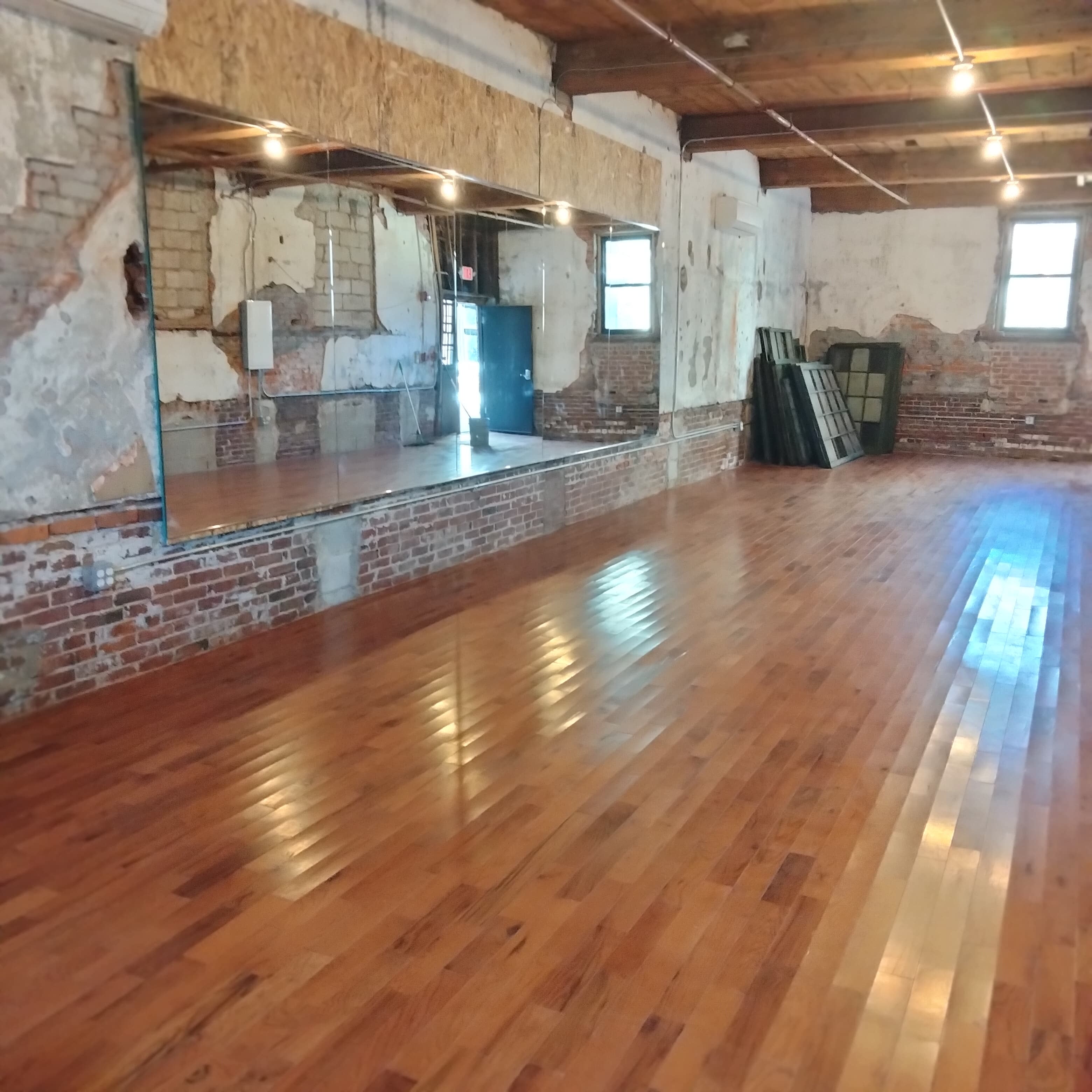 A group of individuals practices dance movements in a spacious studio with wooden floors and exposed brick walls.