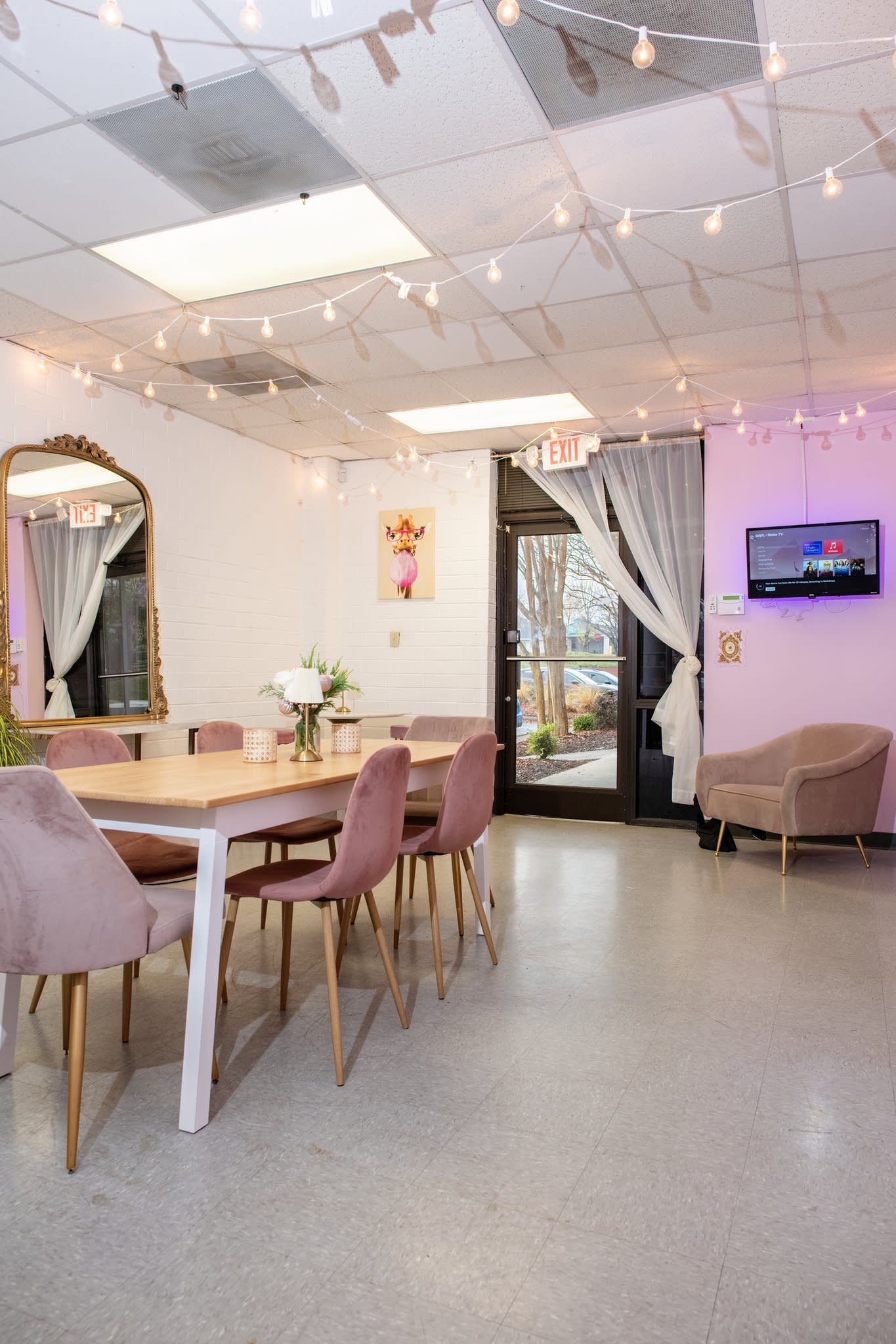 A dinner table is set against a vibrant backdrop of pink flowers and a glowing neon sign that reads "bloom where you are planted."