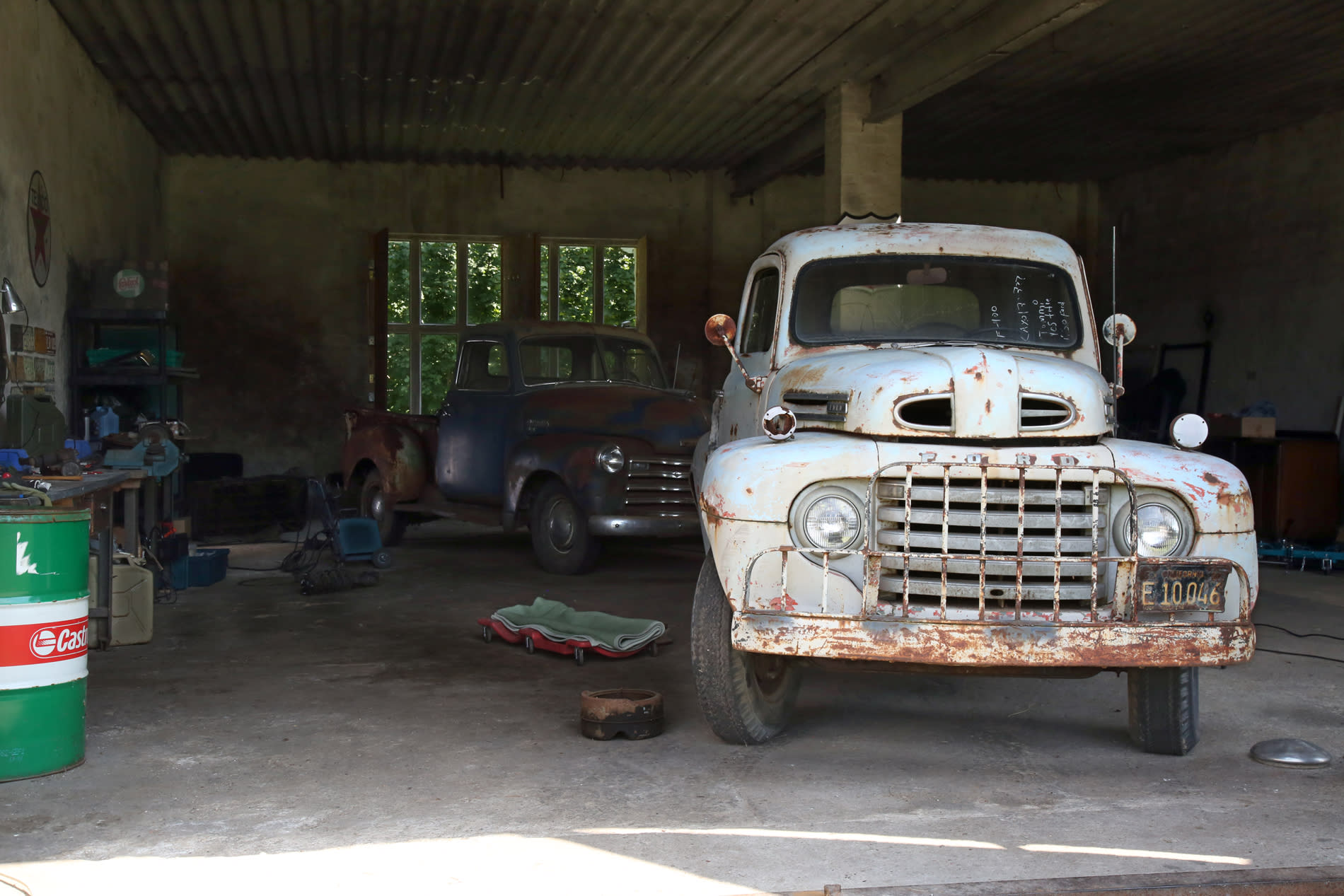 A vintage blue pickup truck is parked inside a rustic garage filled with tools and old car parts.