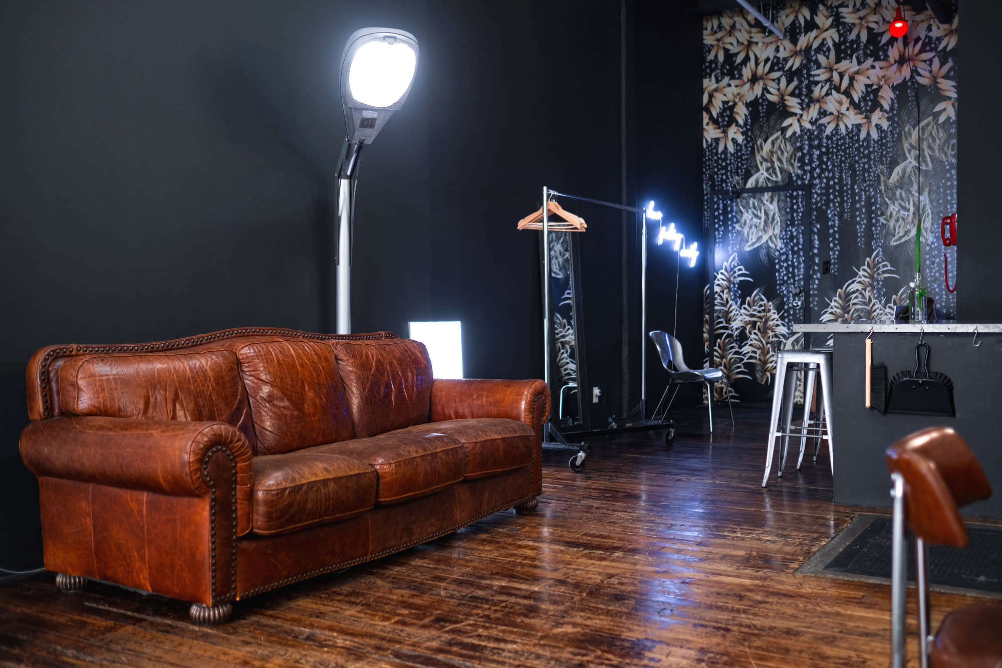 A photo studio setup features a camera on a tripod, a large softbox light, and a reflector, with a wooden table and chair positioned for a shoot.