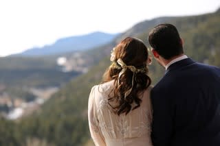 A couple stands hand-in-hand under a floral arch during their outdoor wedding ceremony against a backdrop of mountains.