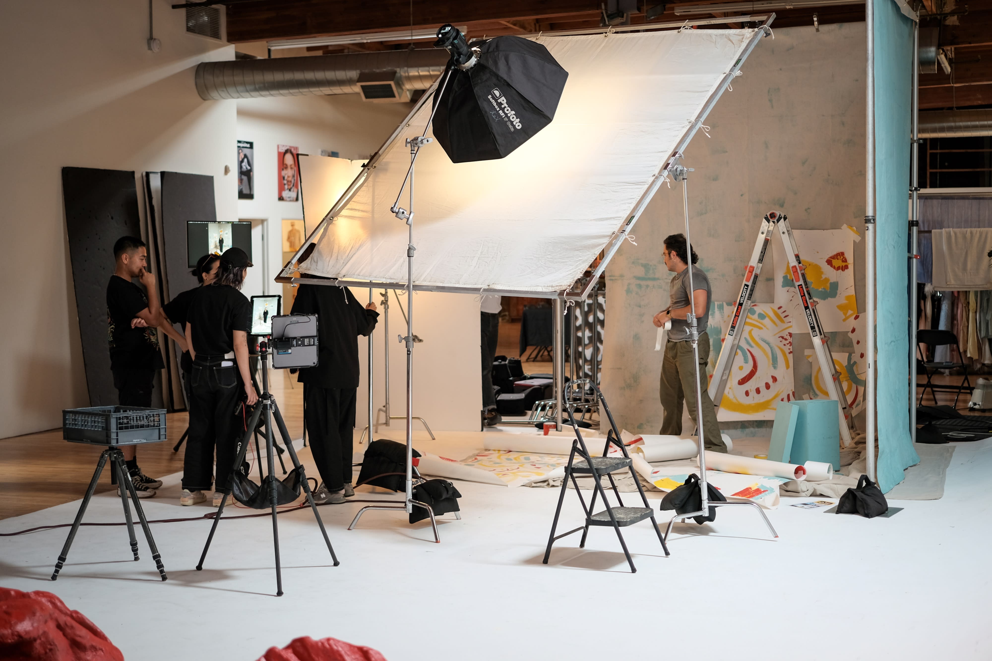 A lifestyle video shoot features a seated interview setup with a person being filmed, surrounded by professional lighting and camera equipment in a spacious studio.