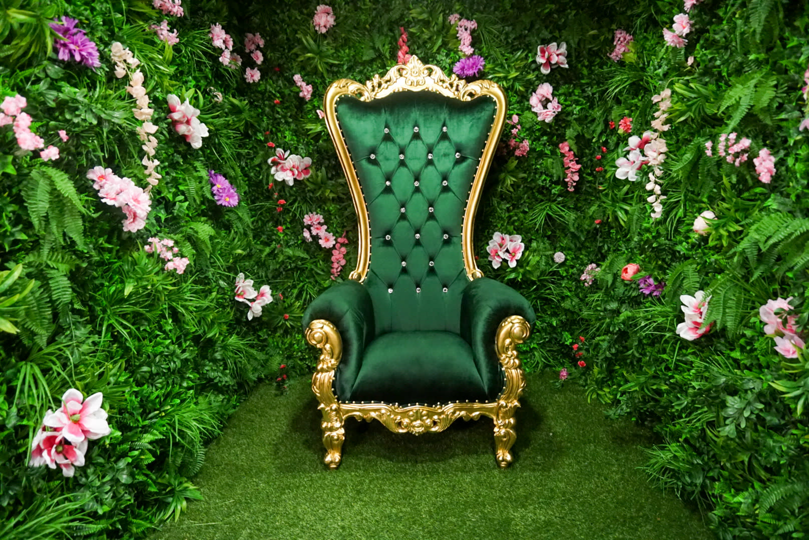 Three individuals pose in elaborate floral attire and grass skirts around a grand, green chair adorned with tropical plants and flowers during a video shoot.