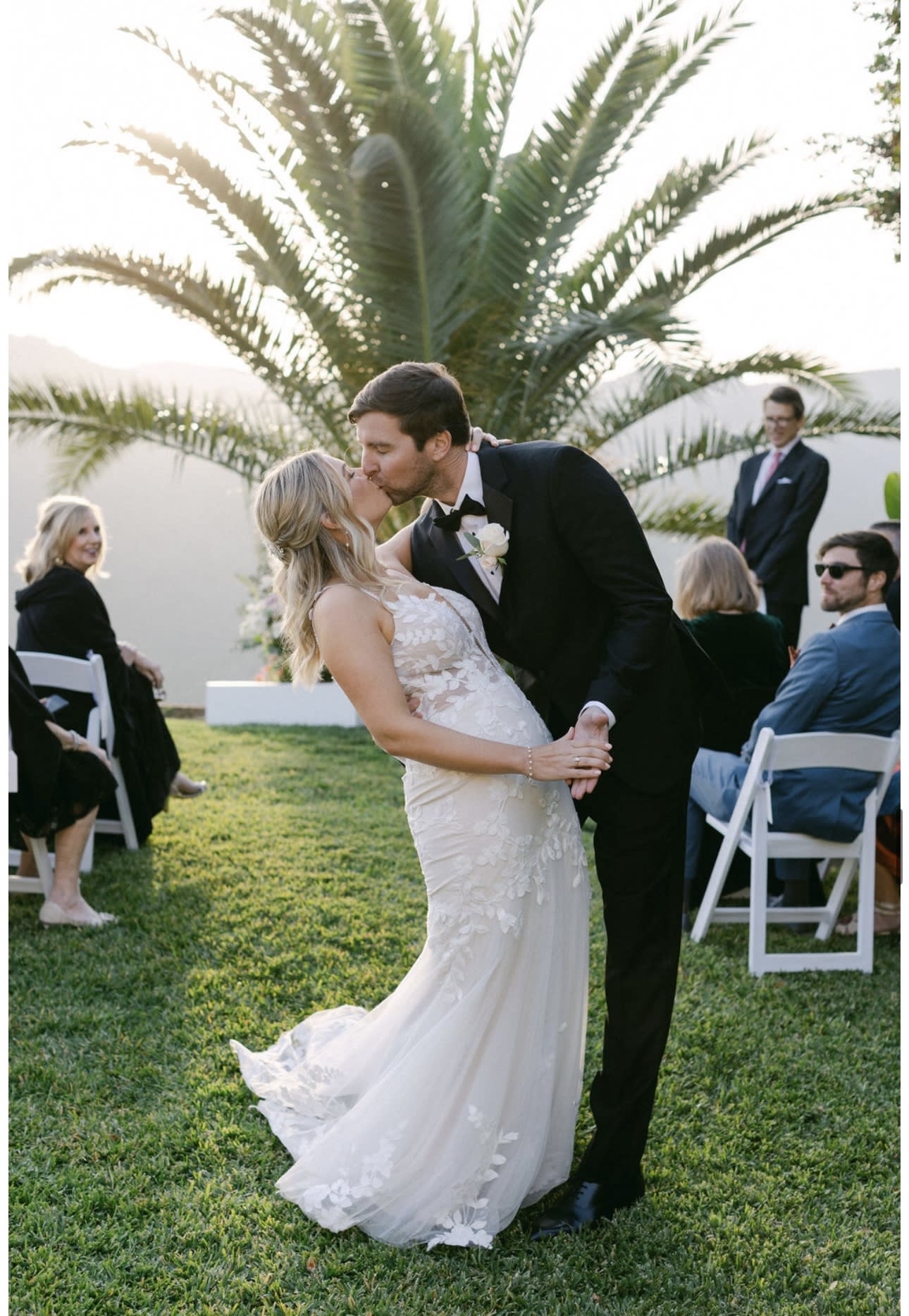 The scene features a beautifully arranged outdoor setting with white chairs facing a large palm tree and floral decorations, overlooking a scenic landscape.