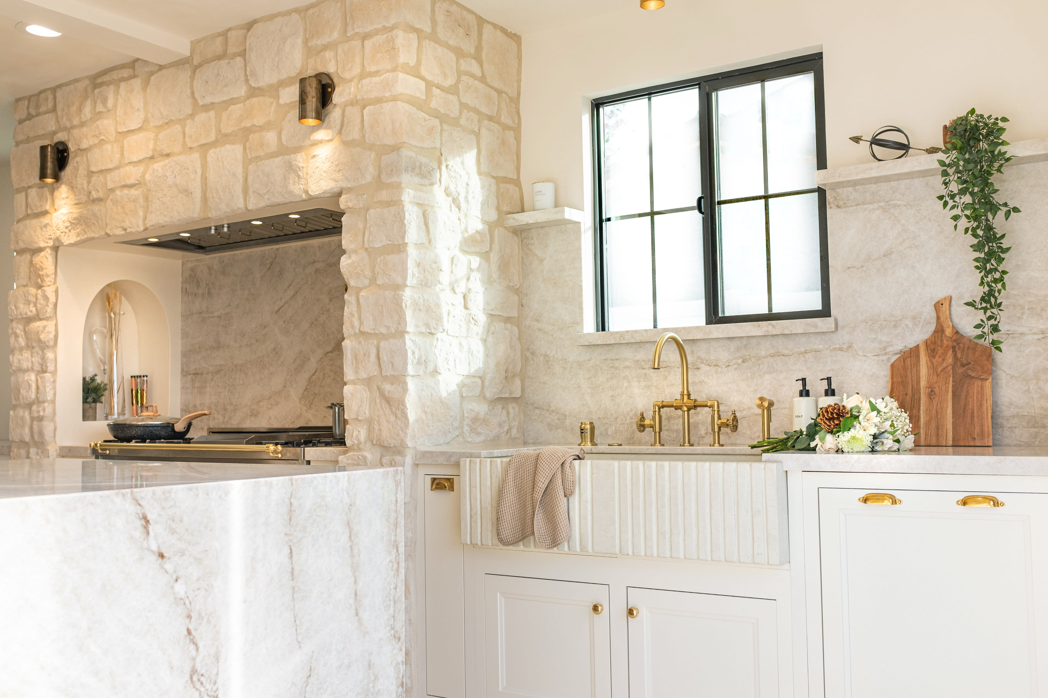 Two women pose in a brightly lit bathroom, one holding a showerhead while the other sits on a ledge, both dressed in stylish cover-ups.