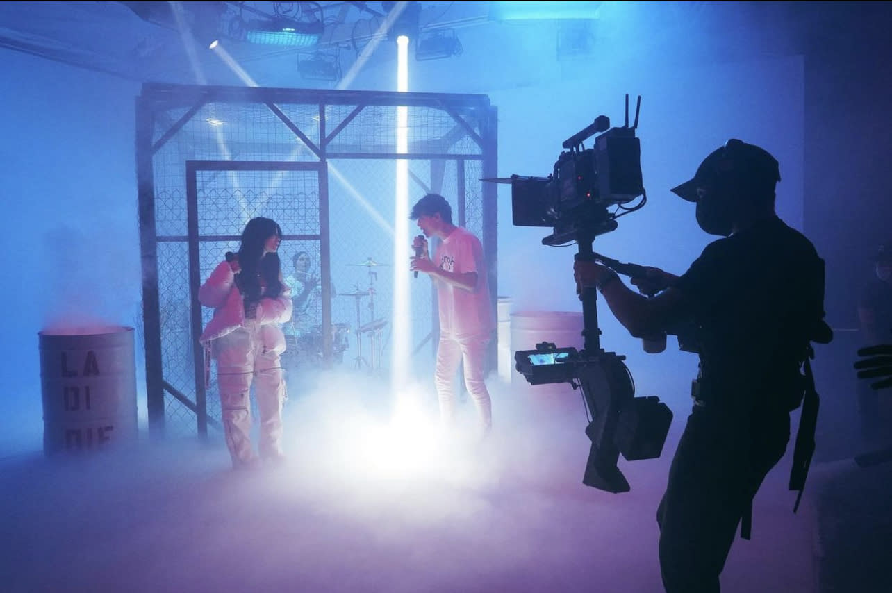 Three individuals are positioned inside a fenced area, engaging in a dynamic scene with weapons drawn in a low-lit industrial setting.