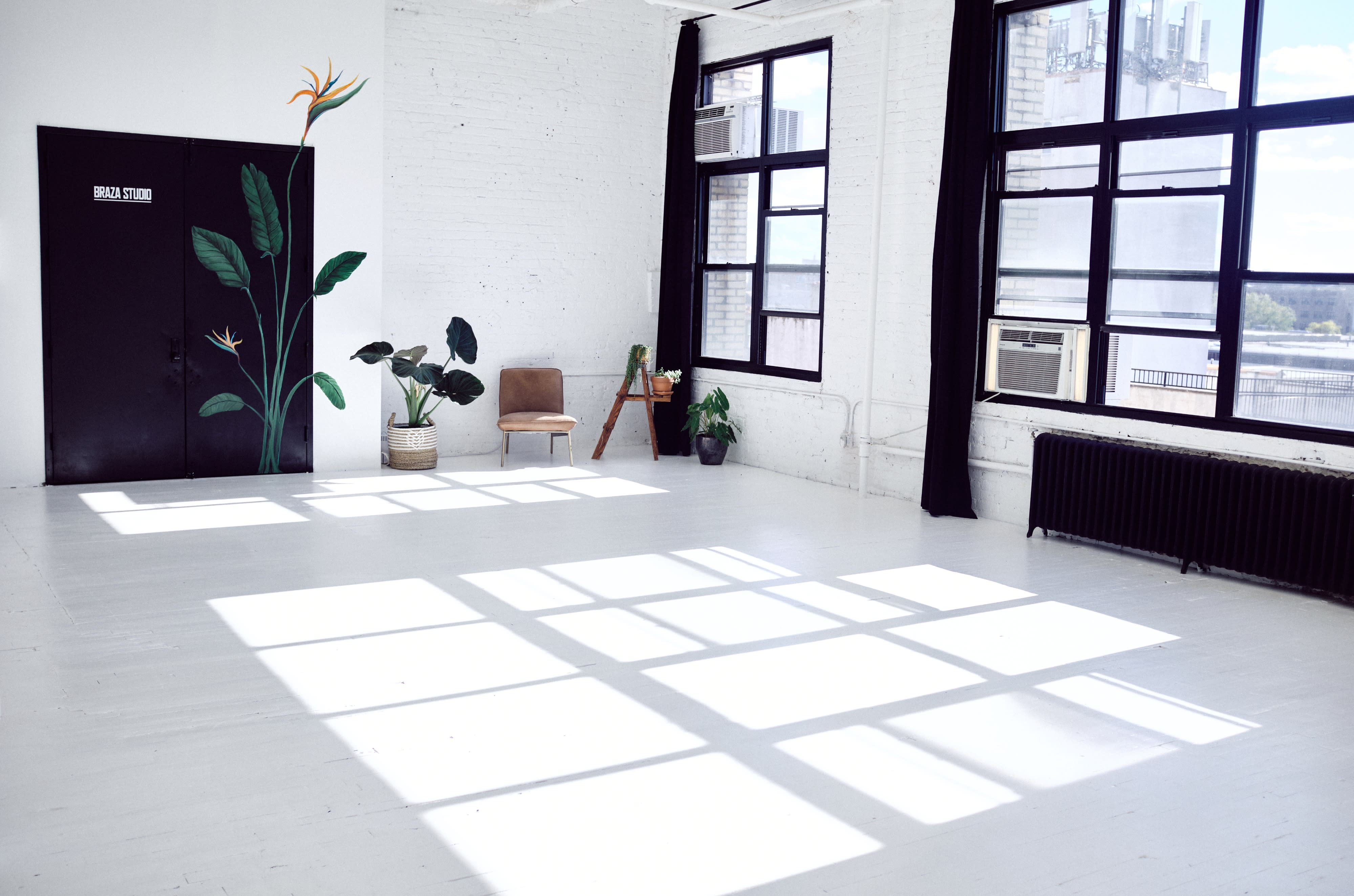 A spacious, empty studio with whitewashed floors and large windows, featuring a few scattered plants and furniture along the walls.