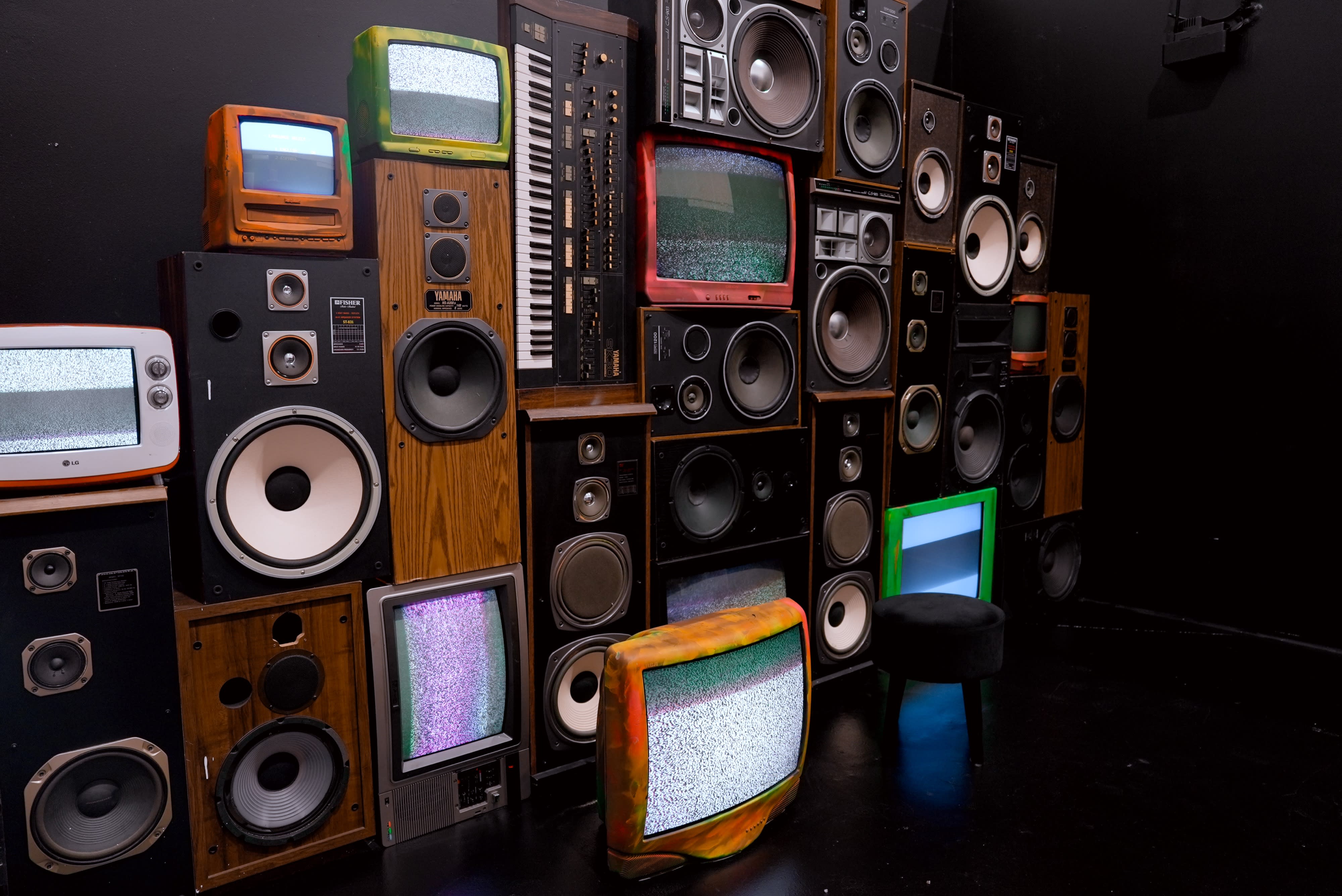 A model poses confidently on a stool in front of a vibrant backdrop of speakers and televisions, wearing sunglasses and fashionable attire.