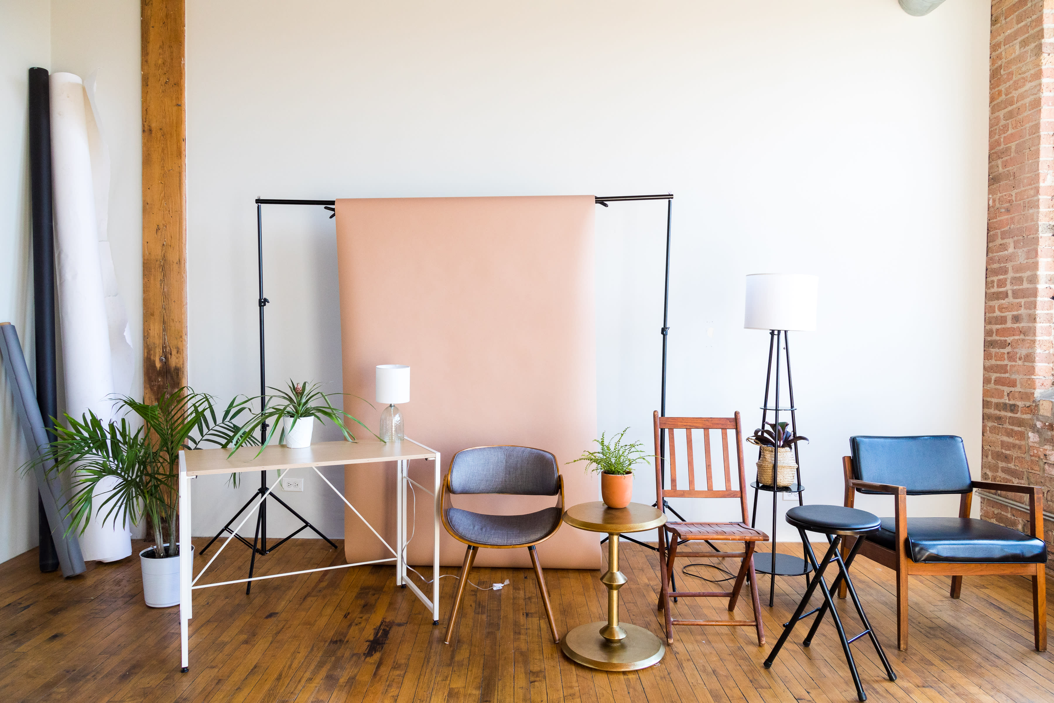 A photoshoot setup features a cozy space with a pink couch, several wooden chairs, and a lamp beside a large window.