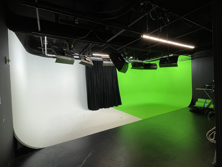 A man sits on a chair in front of a green backdrop while a camera captures the scene.
