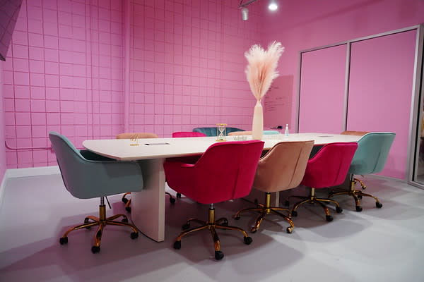 A woman in a pink outfit poses playfully at a desk with a laptop and a poster titled "The Art of Feminism."