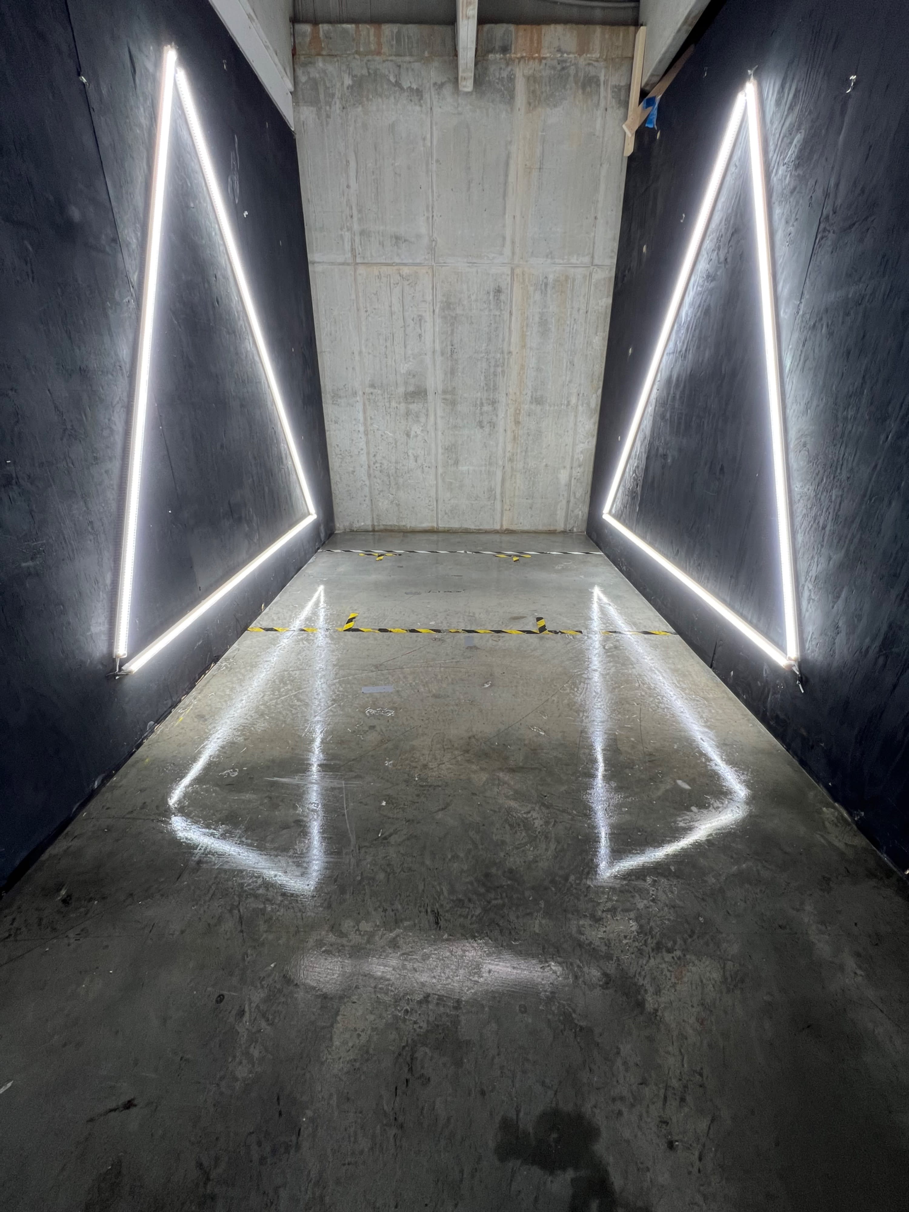A person sits on the floor between two illuminated triangular light fixtures in an industrial space.