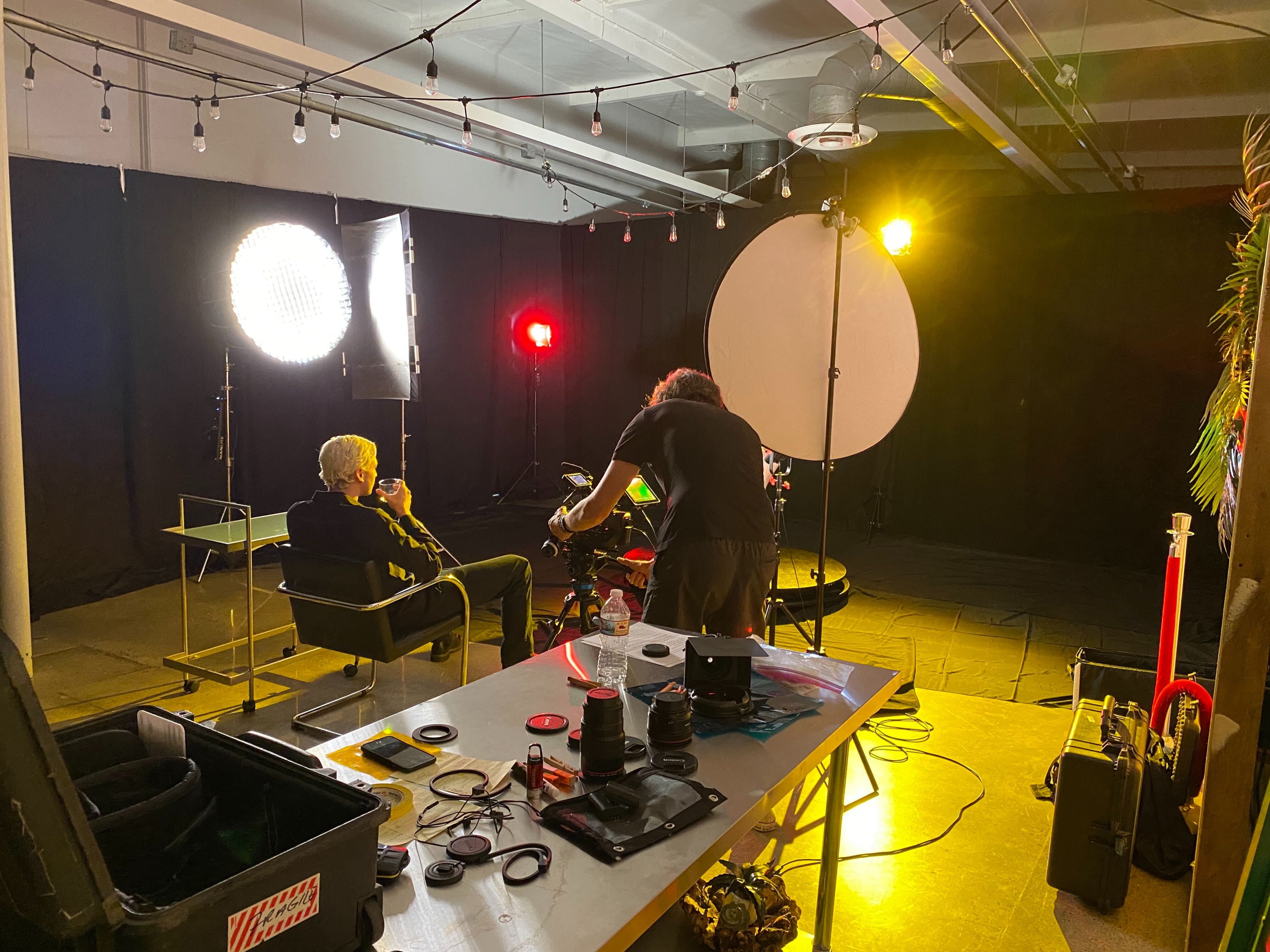 A man sits on a chair in an empty studio, surrounded by multiple cameras and lighting equipment set up for a video production.