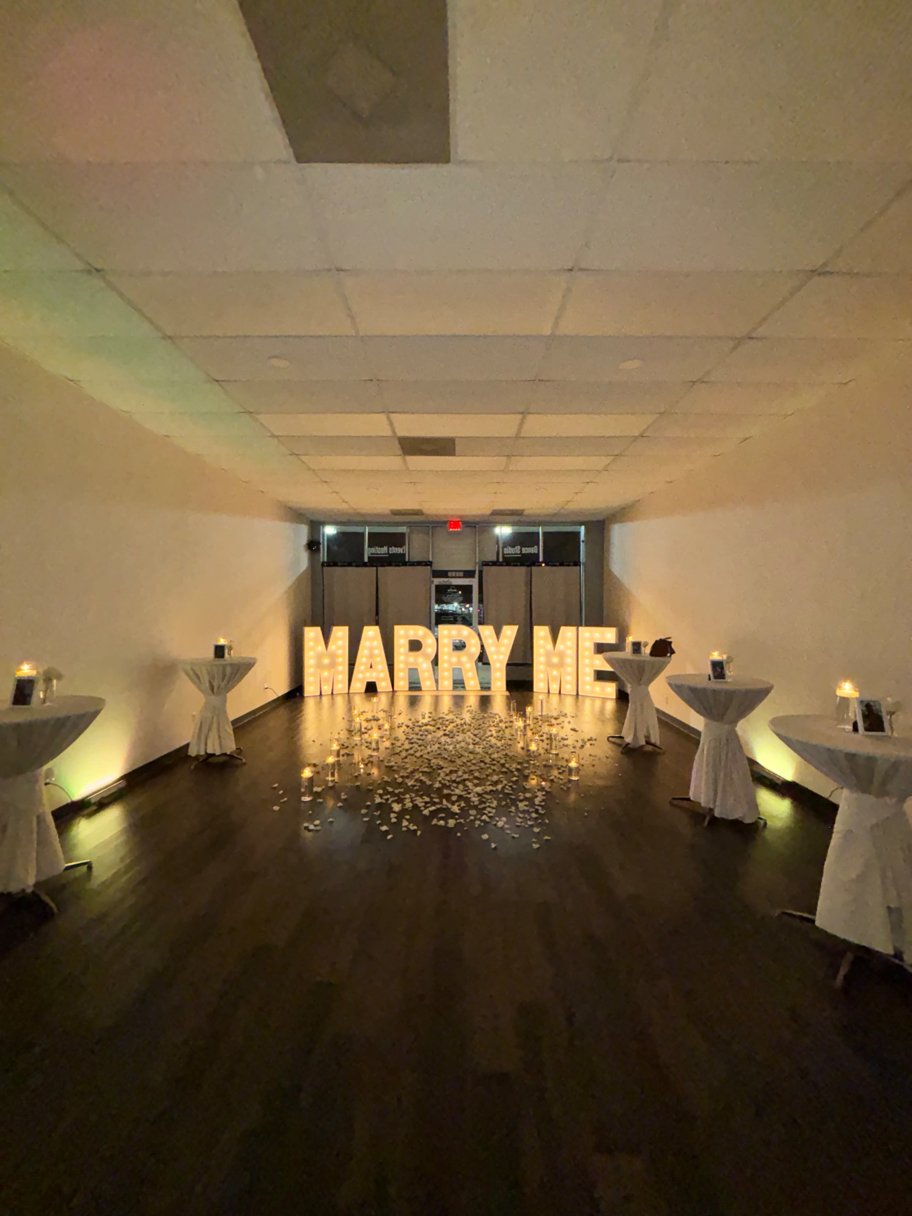 A couple is celebrating their engagement in front of illuminated letters that spell "MARRY ME," surrounded by candles and rose petals on the floor.