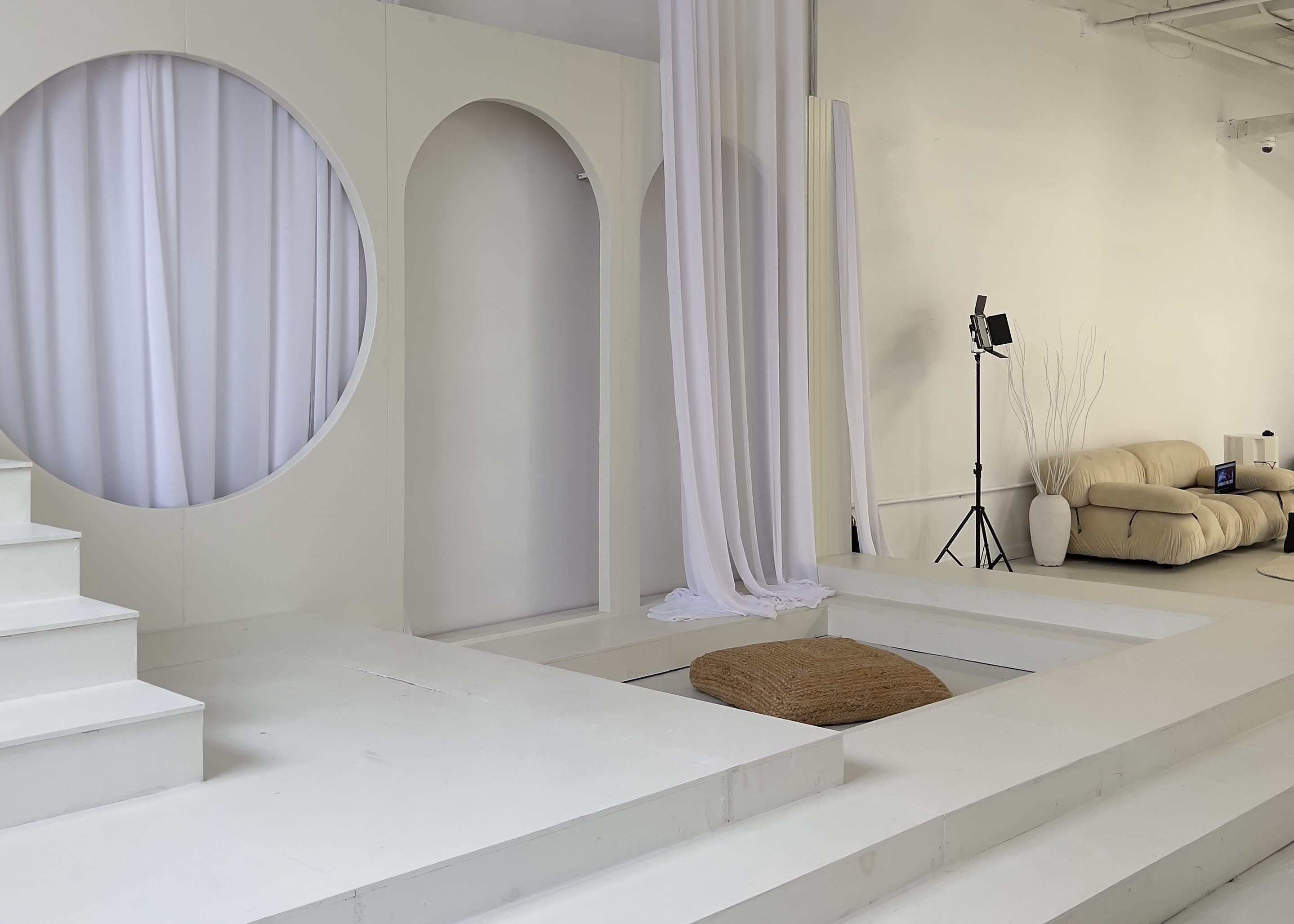 A woman is arranging items on a display stand in a modern, minimalist video shoot set featuring soft arches and a staircase.