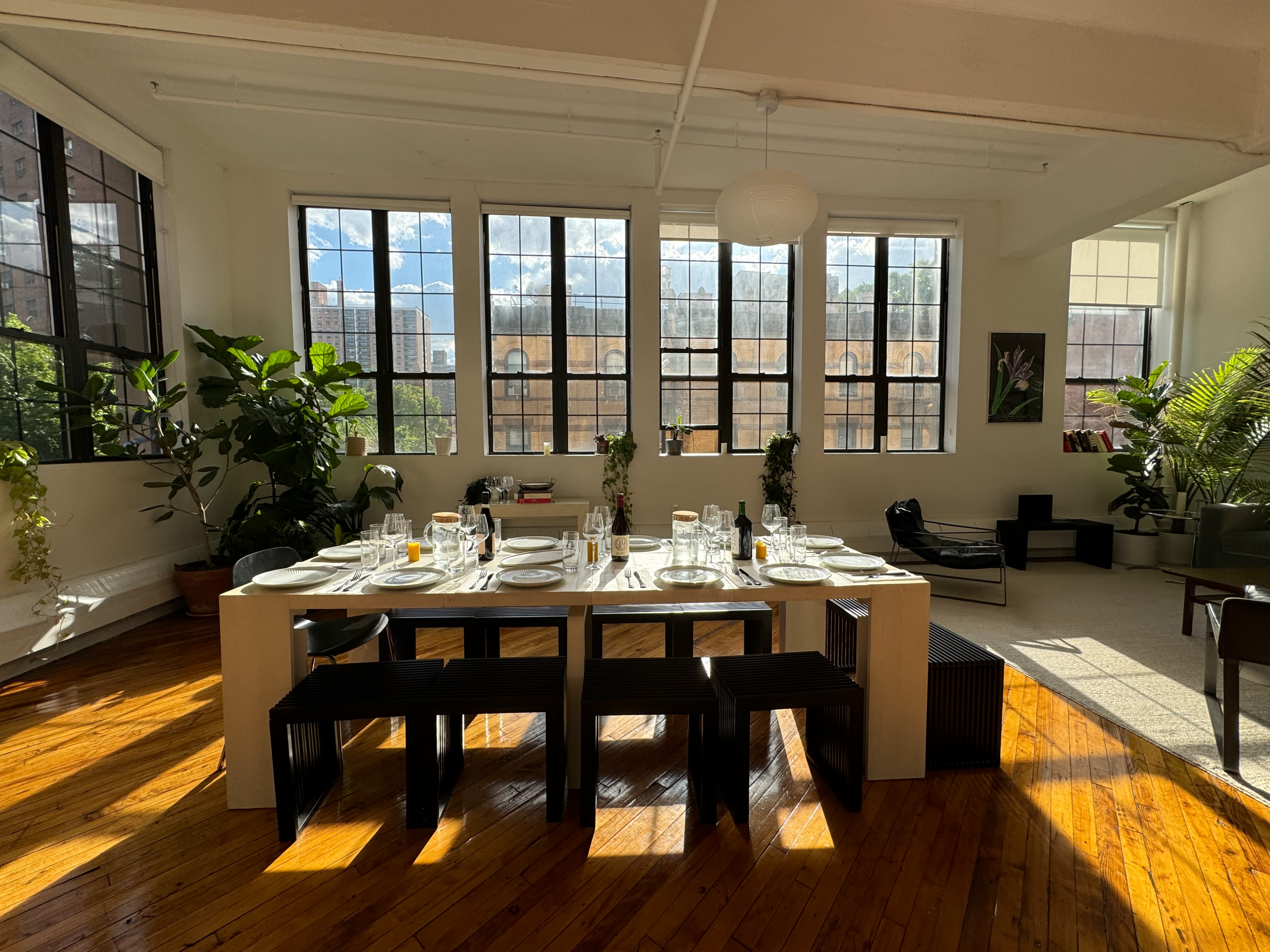 A long dining table is set for dinner in a bright room with large windows, featuring colorful chairs and floral centerpieces.