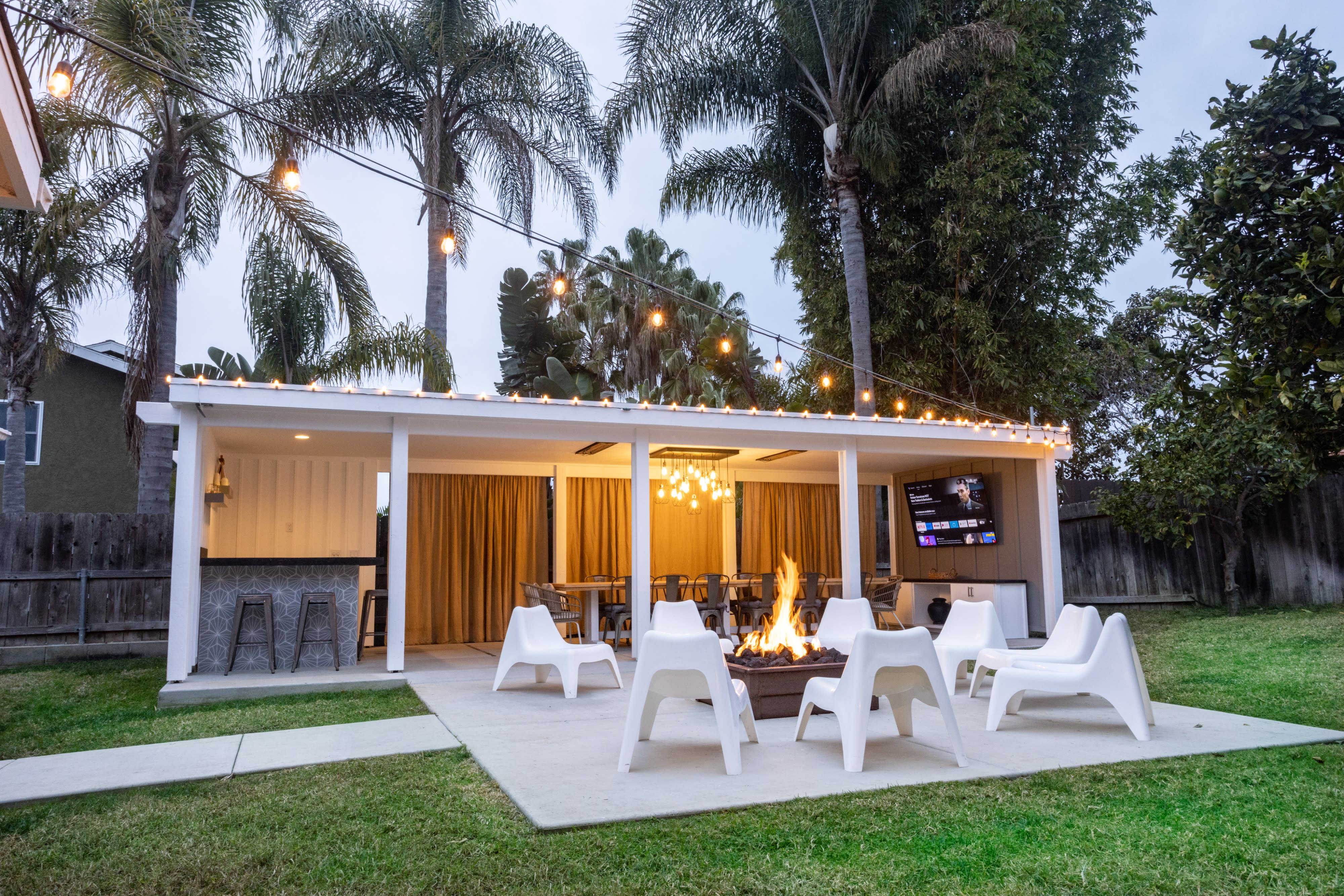 A group of people gathers around a festive outdoor dining setup, with string lights illuminating the space and a fire pit in the center.