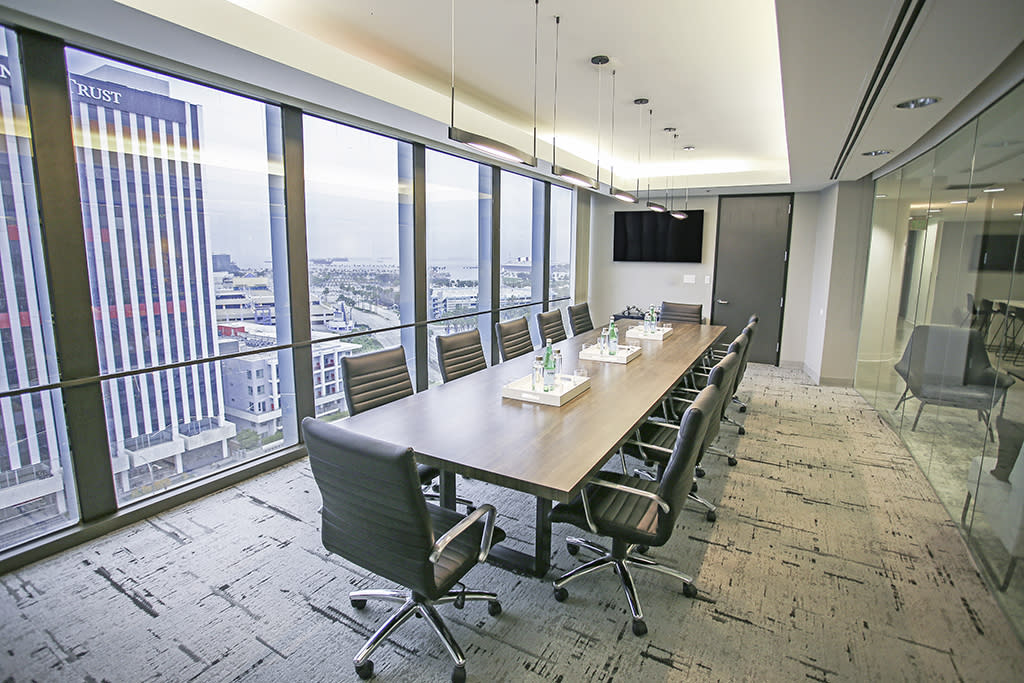 A group of professionals sits around a conference table as a scene is being filmed, with a microphone boom capturing dialogue and cameras recording the action.