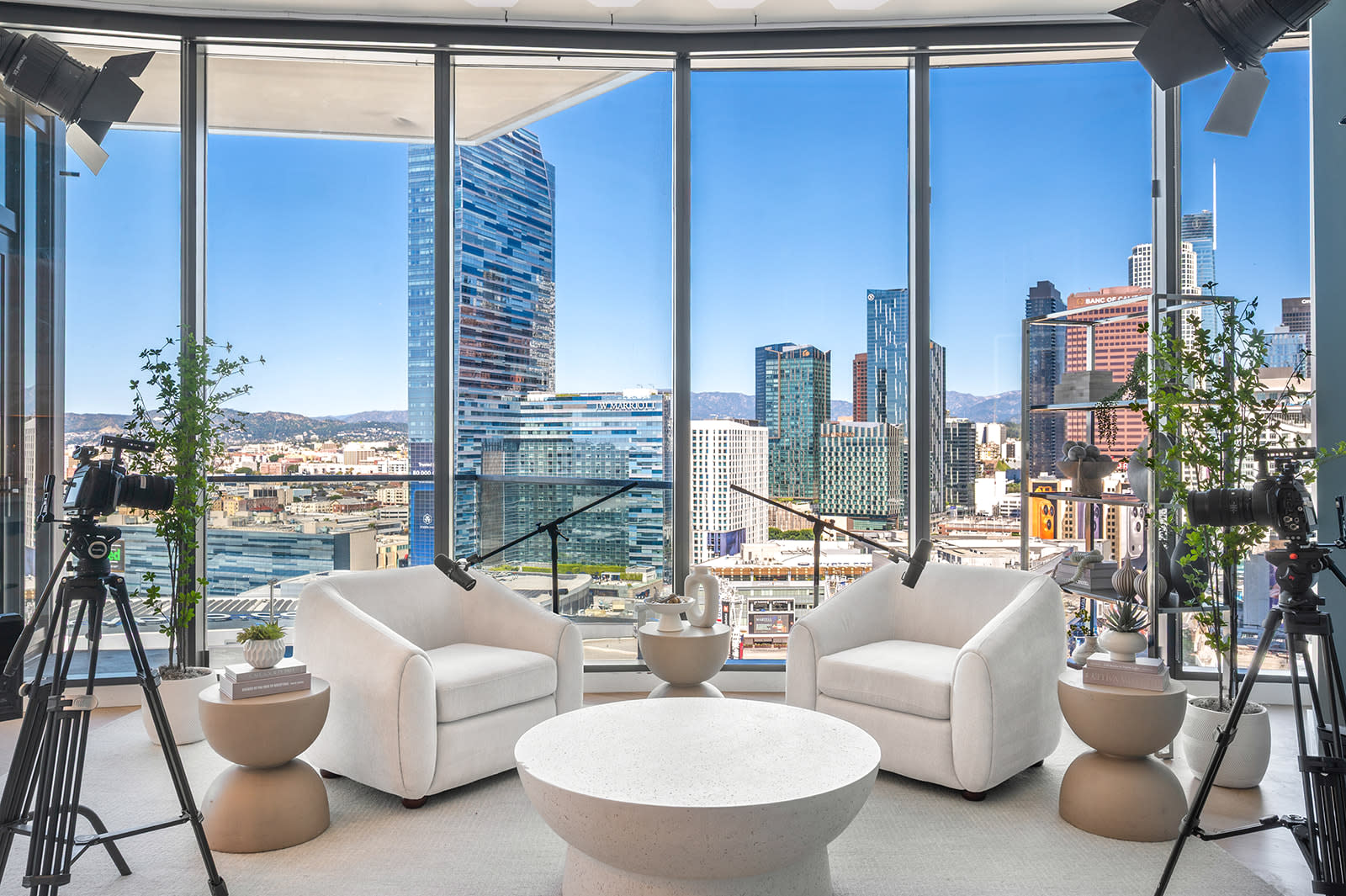 Three individuals are seated on a couch in a modern studio, discussing a podcast while multiple cameras capture the scene against a backdrop of a city skyline.