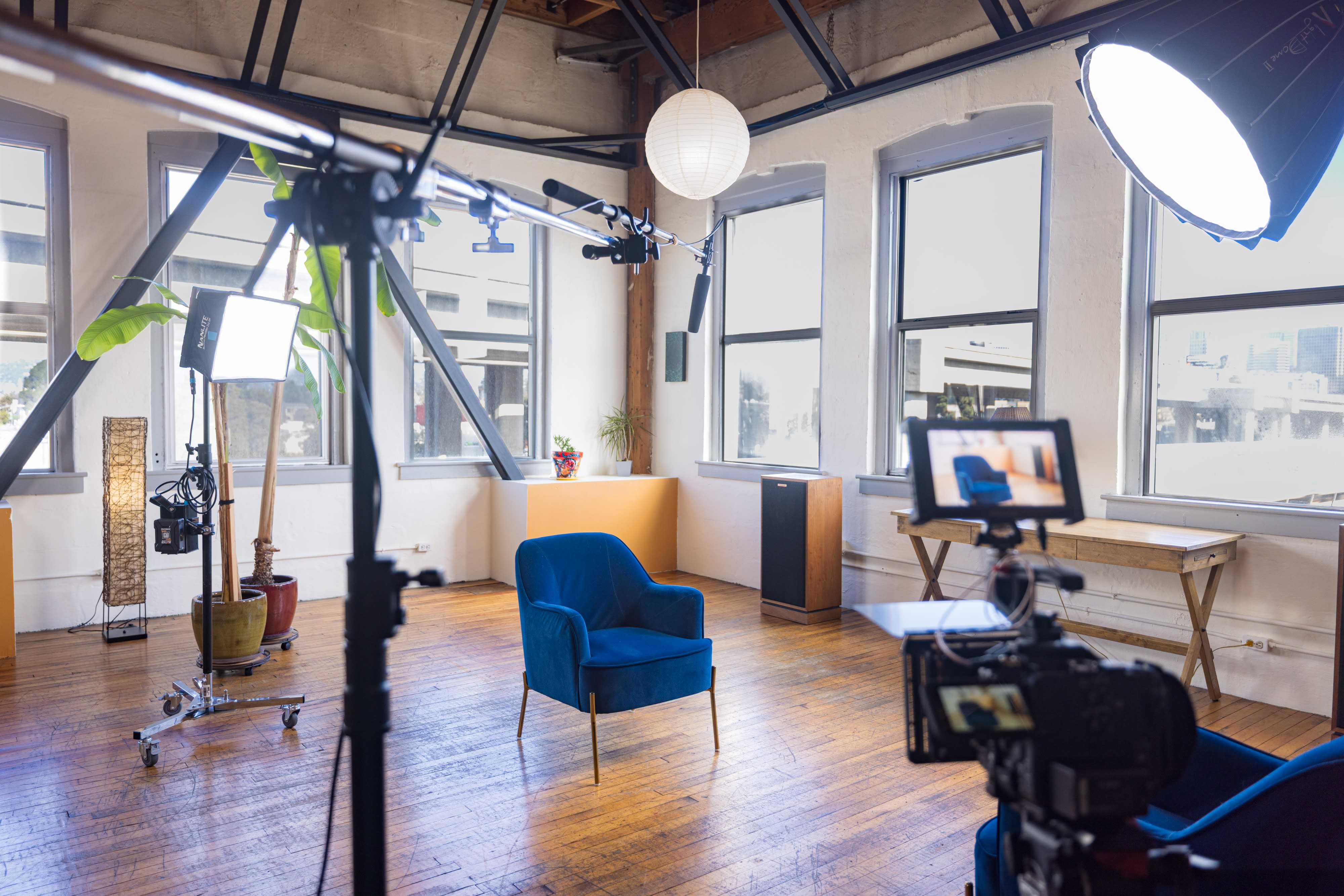 A video shoot is underway in a spacious room, featuring multiple cameras and lighting setups focused on a seated interview subject.