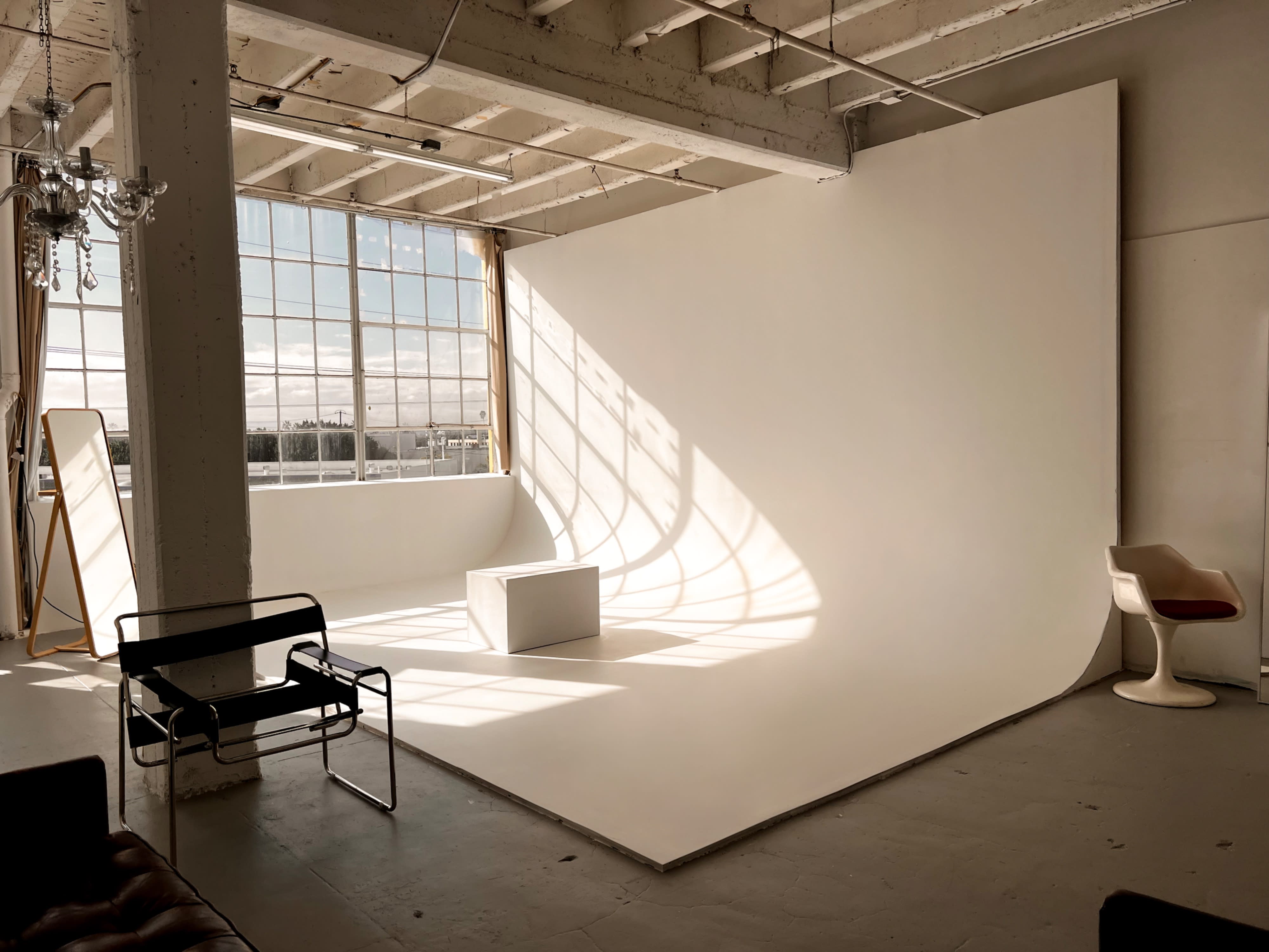 A model poses on her knees beside a modern chair while holding a phone in a minimalist studio setting.