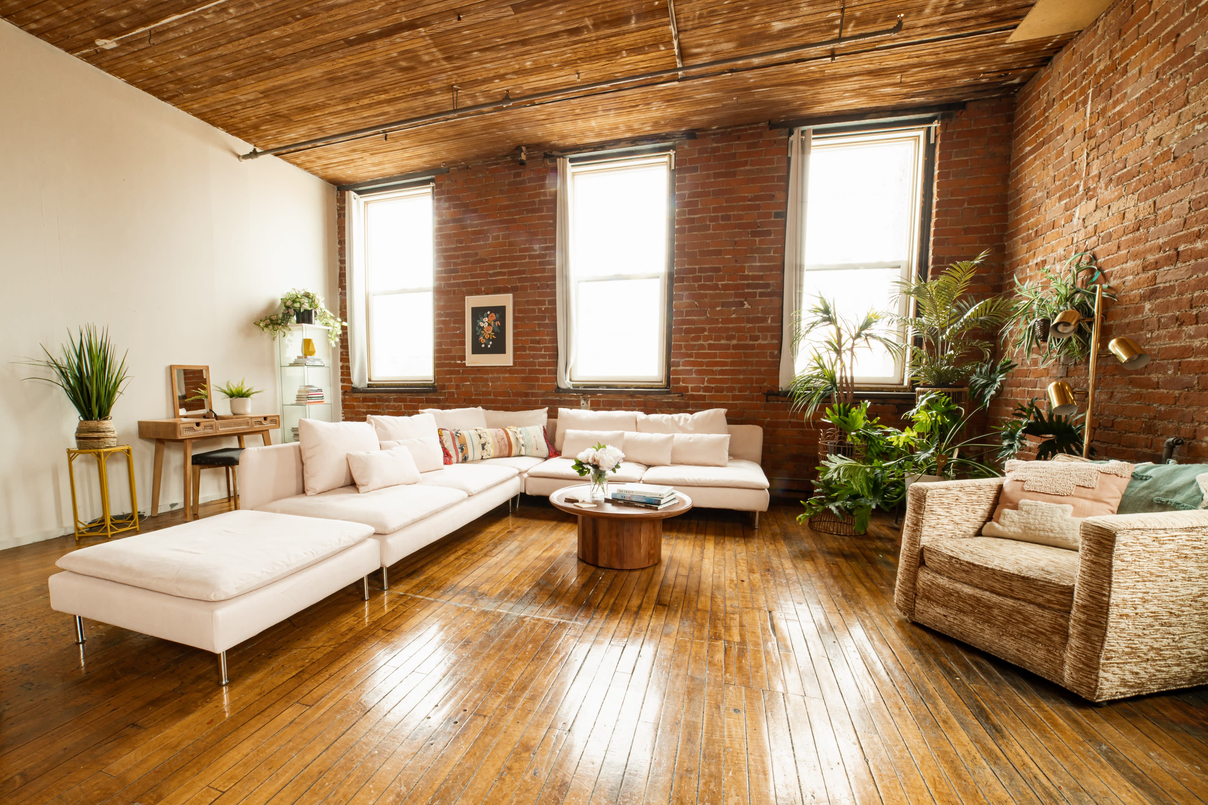 A model poses on a leather couch in a relaxed position, wearing stylish attire and black boots, set against a backdrop of exposed brick and plants.
