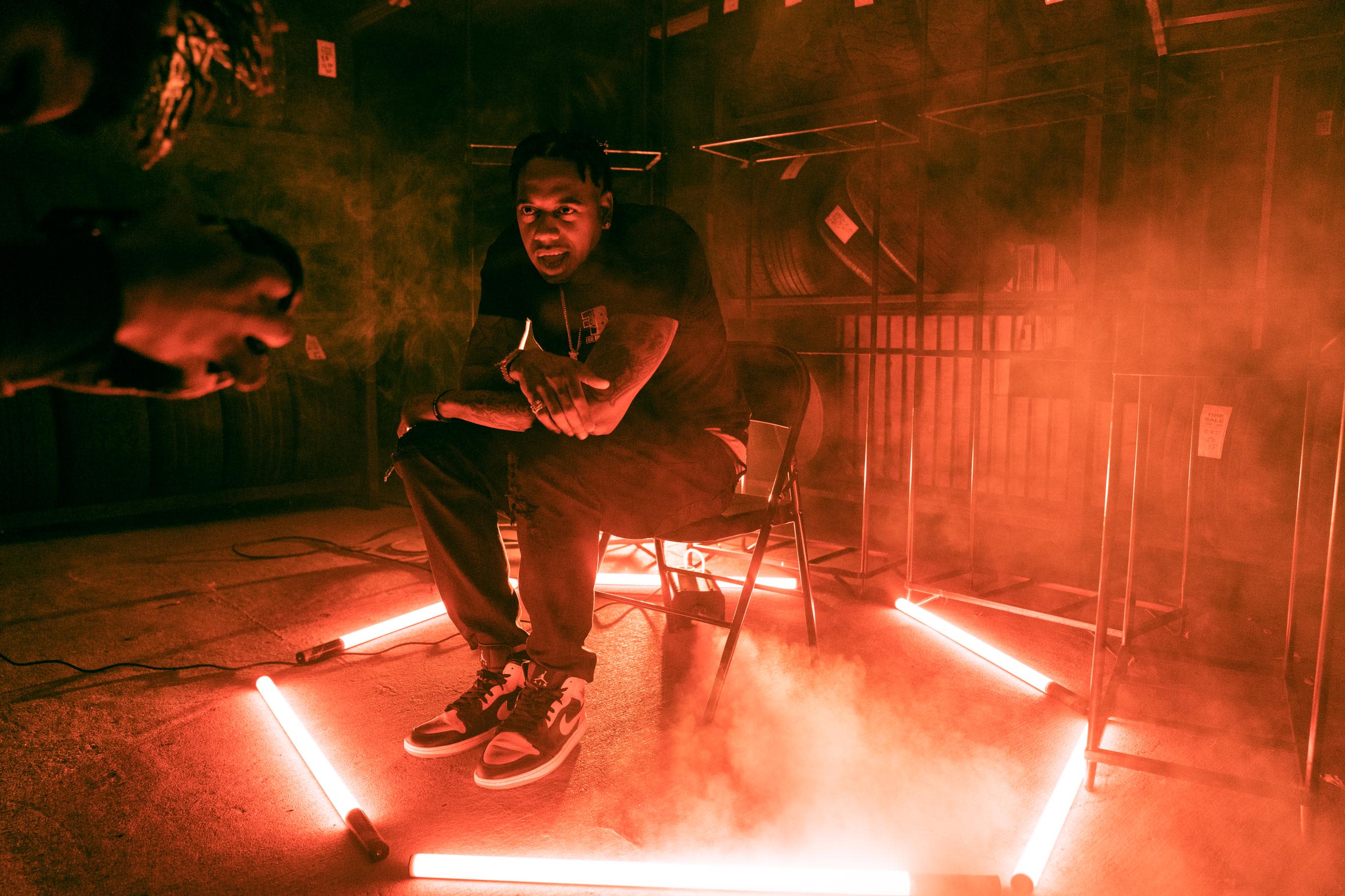 A person is crouched on the ground in an industrial space surrounded by stacks of tires, with smoke billowing above.