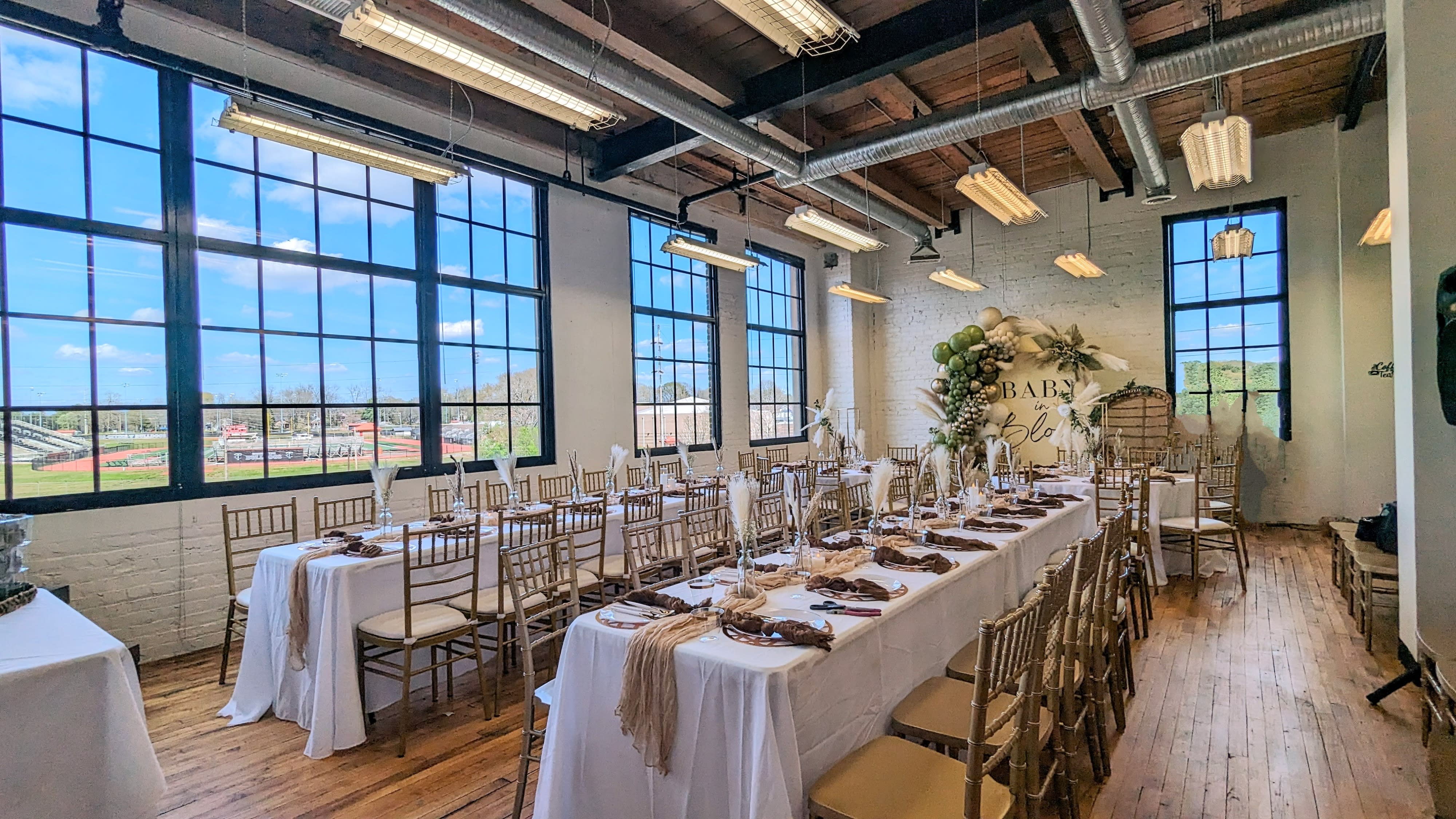 A decorated banquet hall features a long table set for a baby shower, complete with elegant tableware and a backdrop displaying "BABY in BLOOM."
