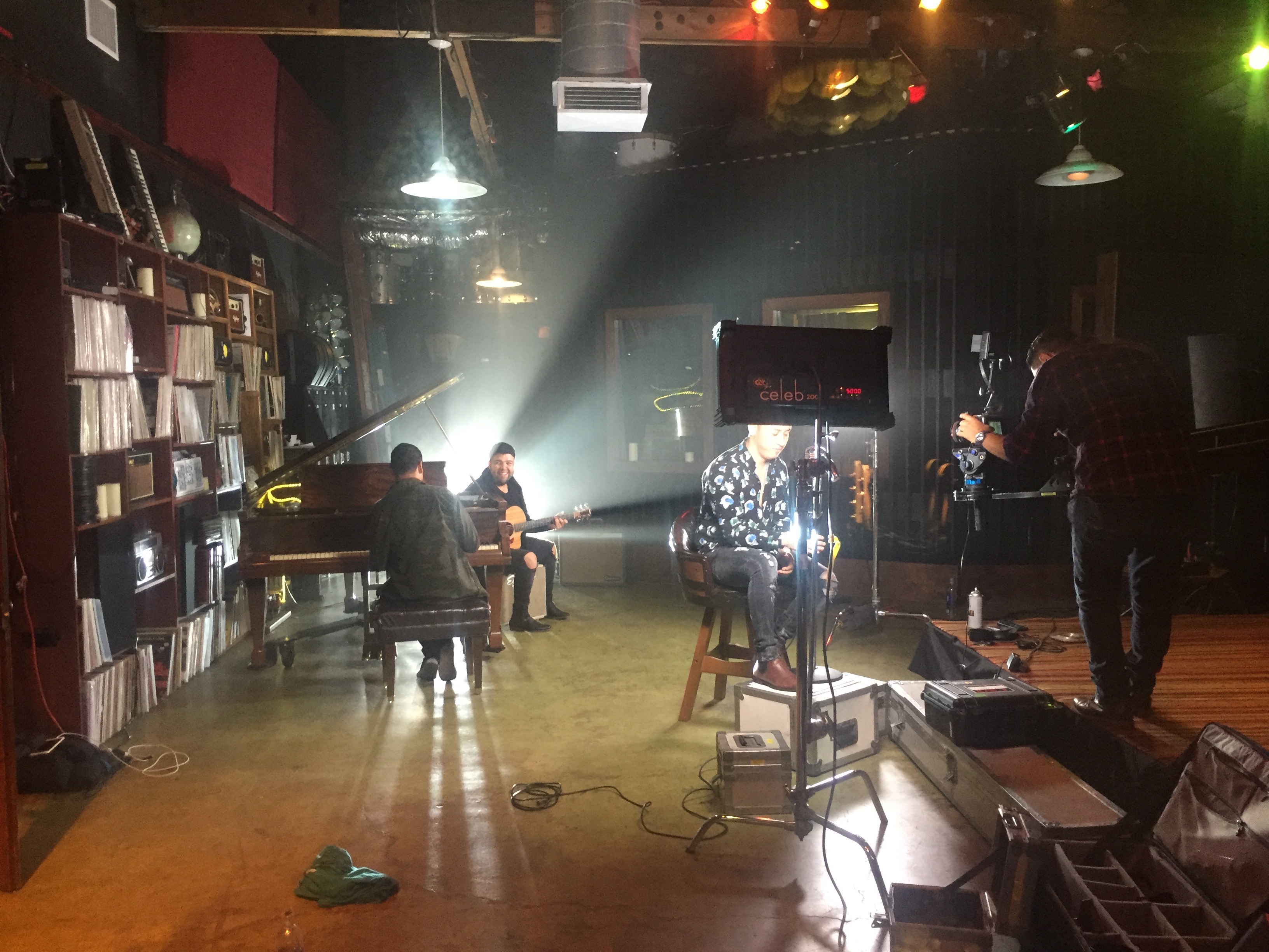 A musician sits on a stool in a recording studio while another person plays a keyboard in the background.