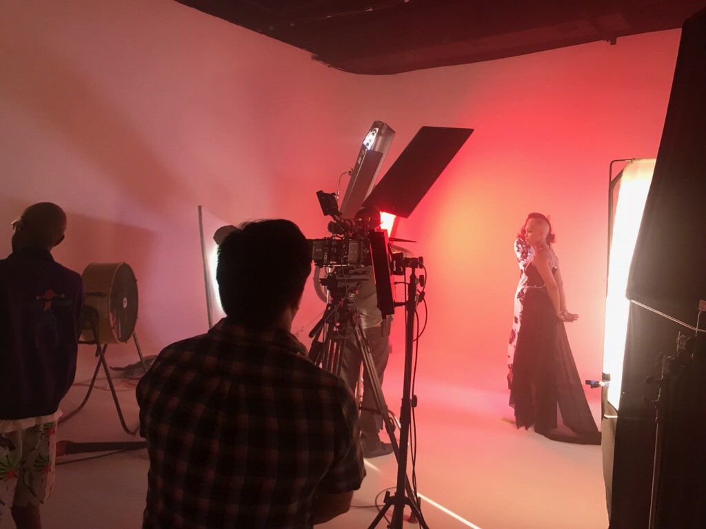 A person poses with their hands behind their back while another individual takes photographs under a large umbrella light in a studio.