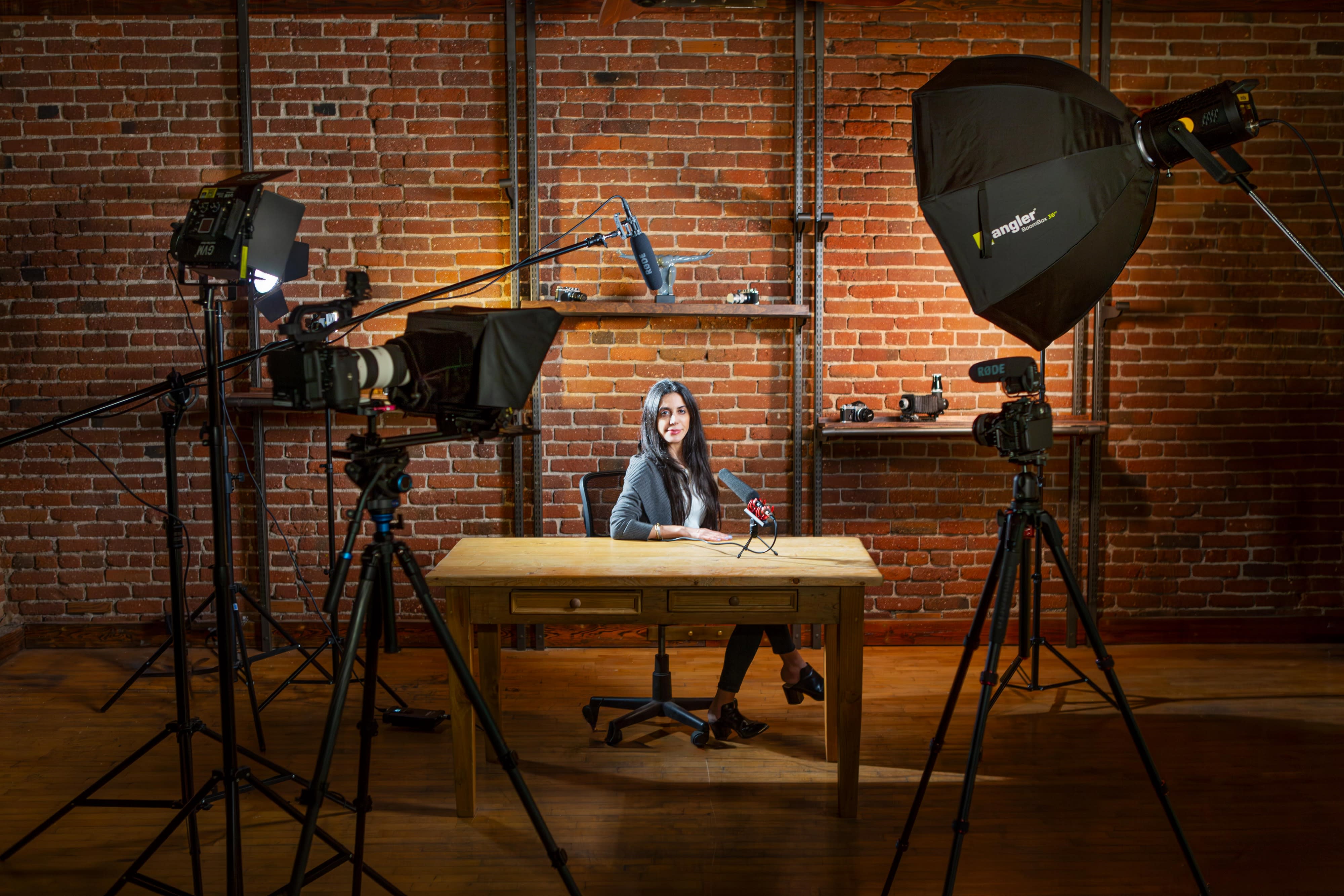The scene captures a photoshoot setup with a subject sitting at a table, illuminated by multiple studio lights in a dimly lit space.