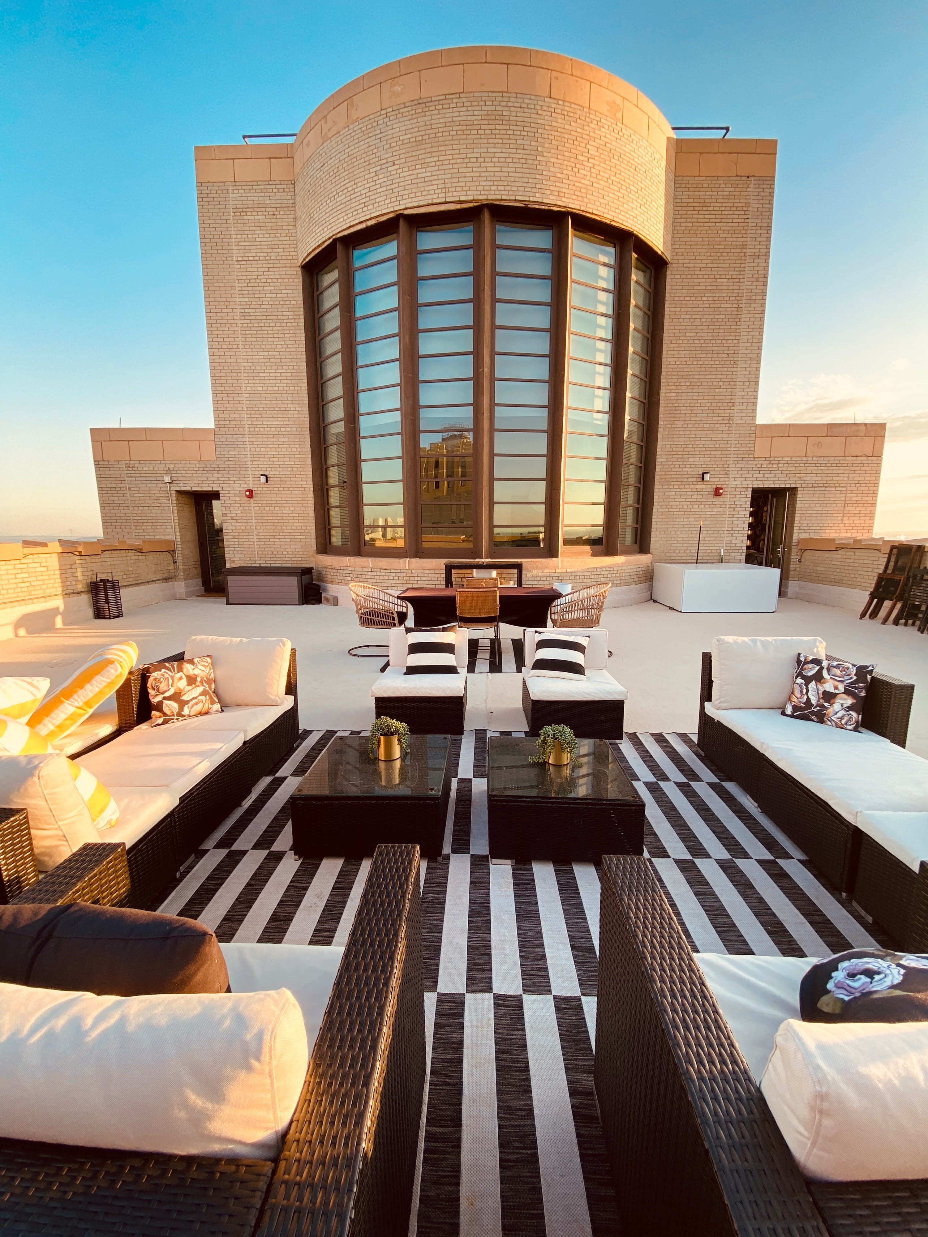 Two people are having a conversation on a rooftop patio with a grill, overlooking a city skyline at sunset.