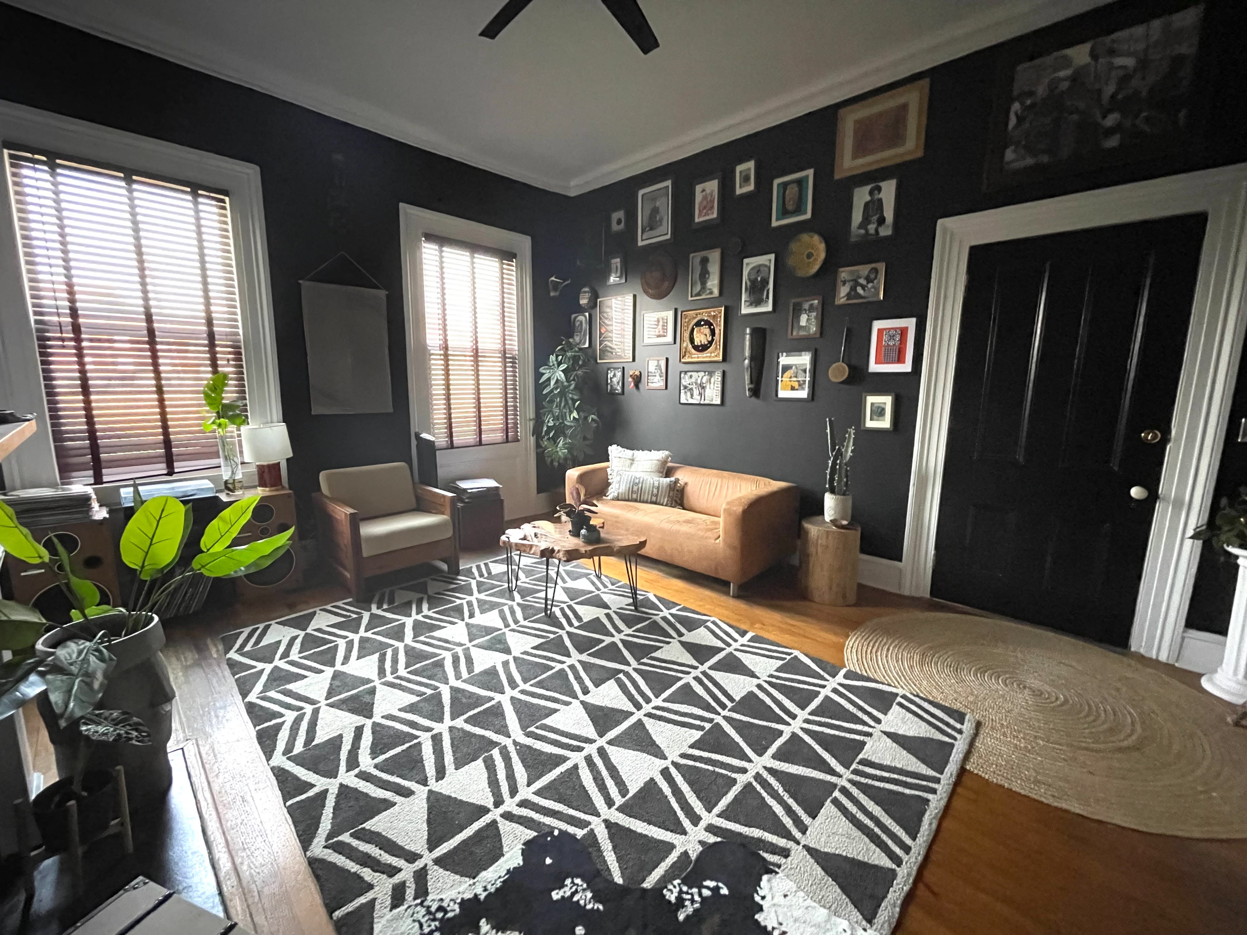 A cozy photo shoot features three candles surrounded by dried leaves and a pinecone on a wooden table, with framed artworks on the dark wall behind.