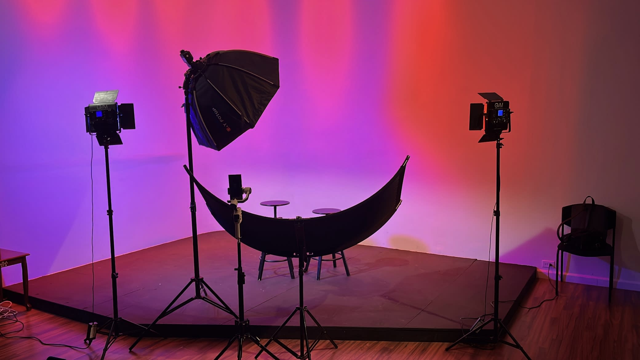 A woman sits confidently on a director's chair, holding a microphone, in a well-lit studio with purple lighting.