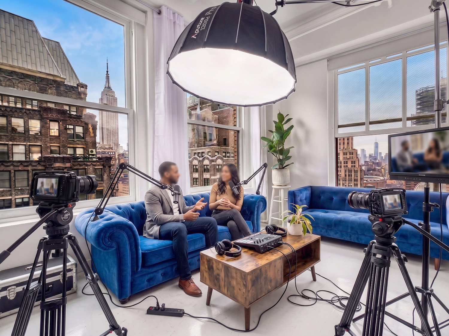 A photo shoot takes place in a well-lit studio featuring a couch and chairs, with large windows showcasing a view of the Empire State Building.