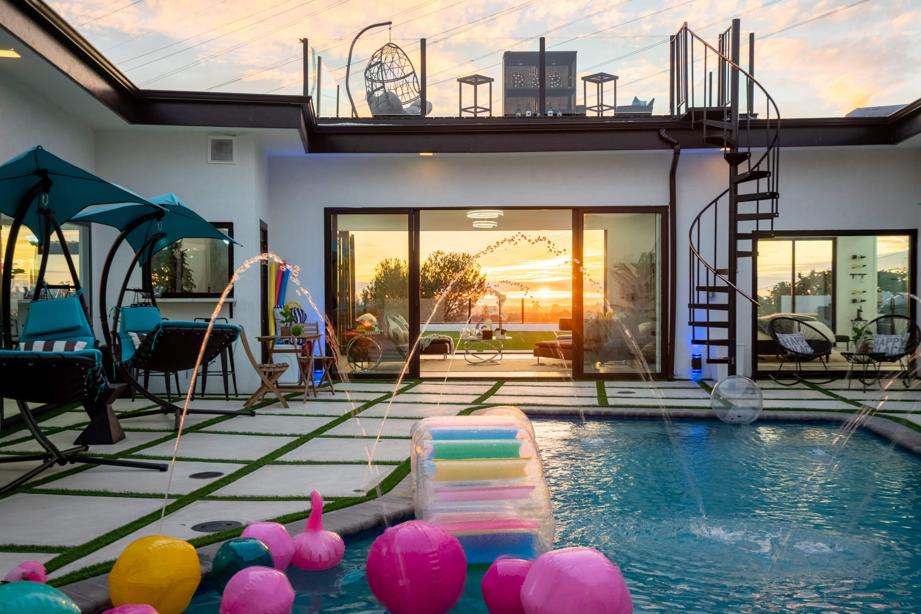 A group of people gathers around a swimming pool with colorful floaties in a modern outdoor setting.