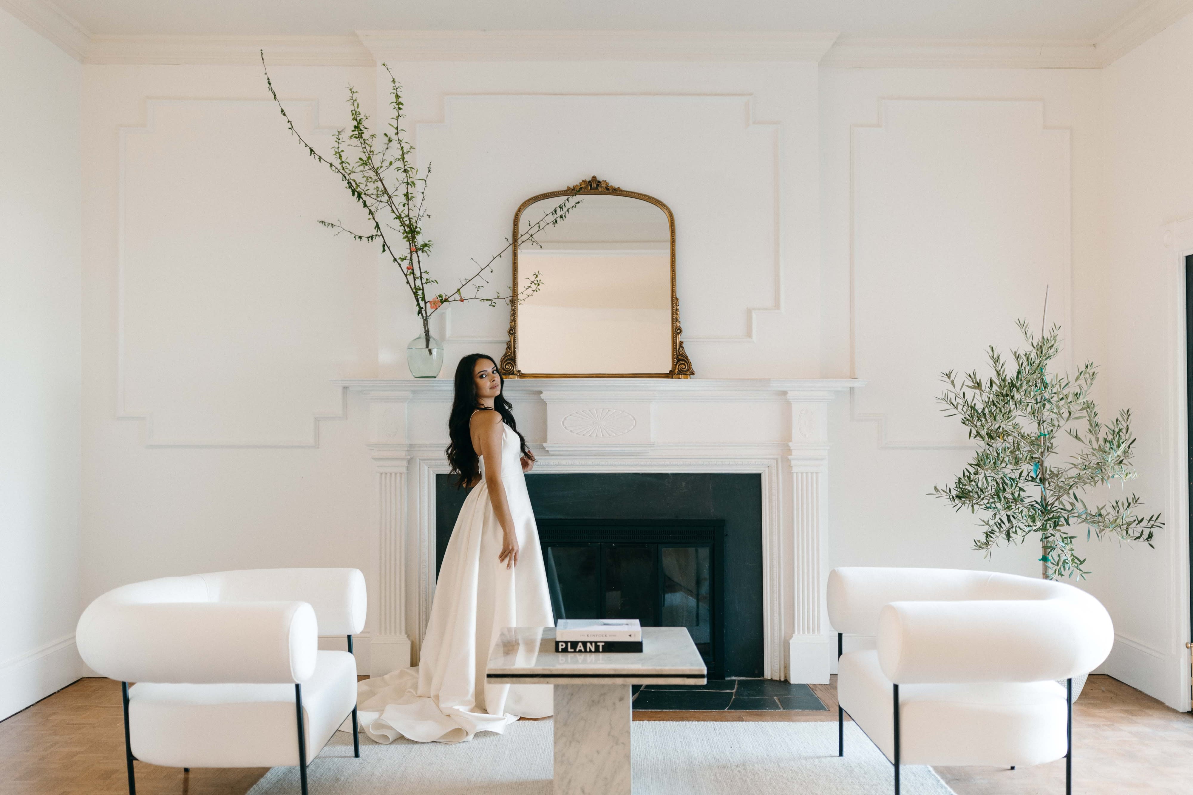 A woman in a flowing white gown stands by a large window, gazing outside.