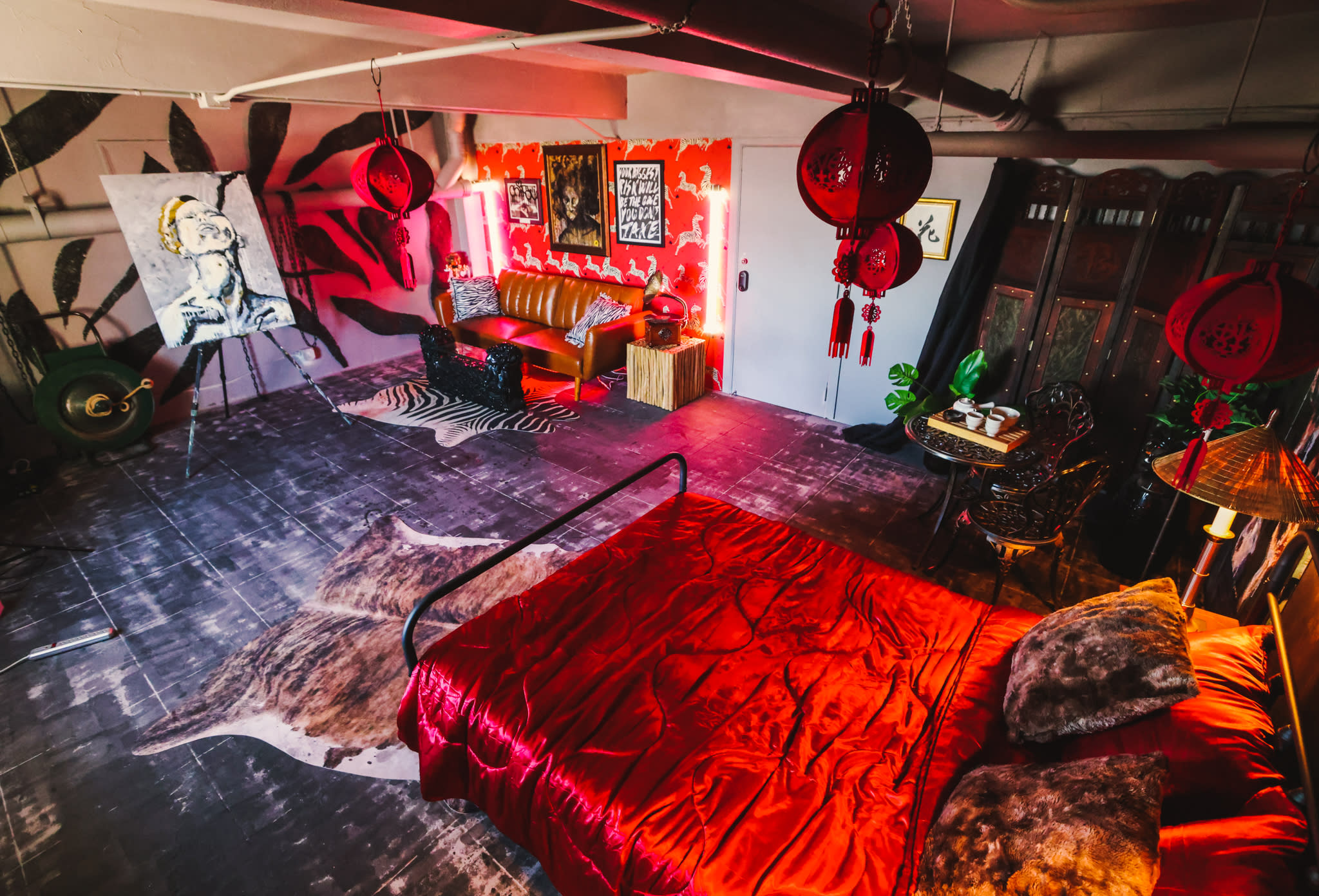 A person poses on a red bedspread in a dimly lit room adorned with candles and an ornamental fan.