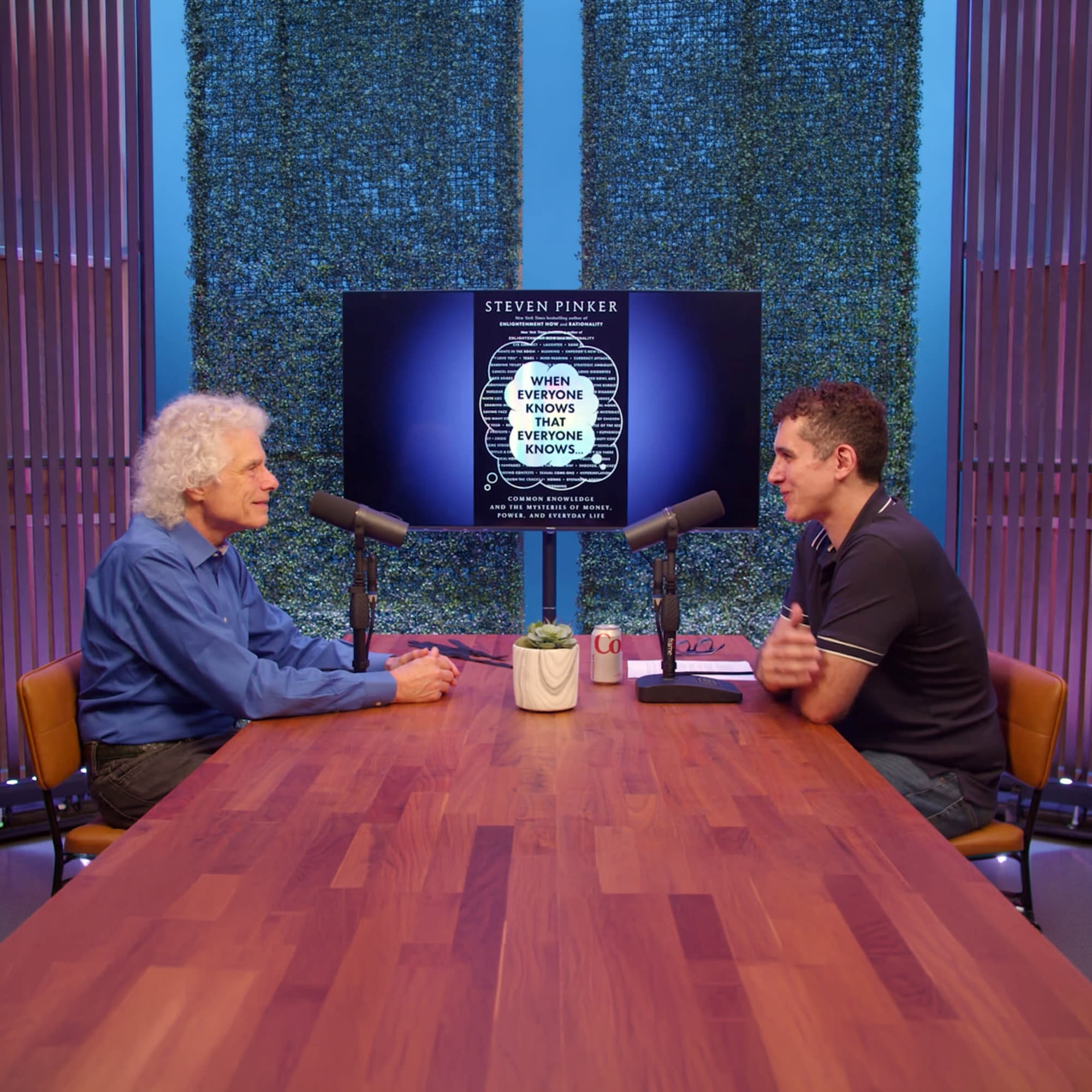 Two hosts sit on armchairs in a studio setting, preparing for a podcast video shoot with microphones in front of them and a simple table between them.